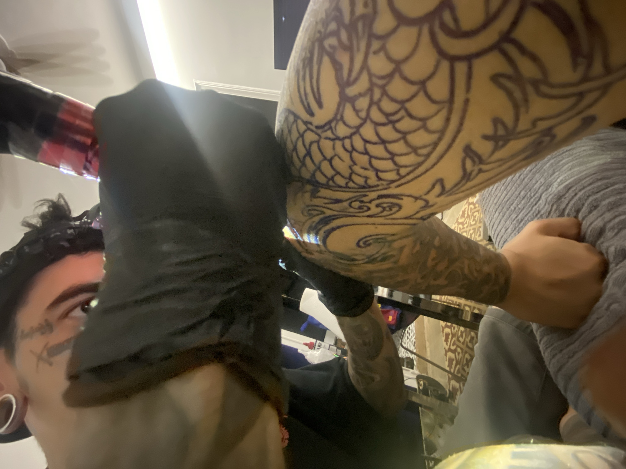 A person with facial tattoos and gauges in their earlobes, wearing a black glove, appears to be tattooing a dragon scale design on another person's arm. The tattoo artist is holding a tattoo machine. The person receiving the tattoo is wearing a gray shirt. The location is likely in Barcelona, Spain, based on the presence of Spanish-language text and potentially a Spanish flag in the blurry background.