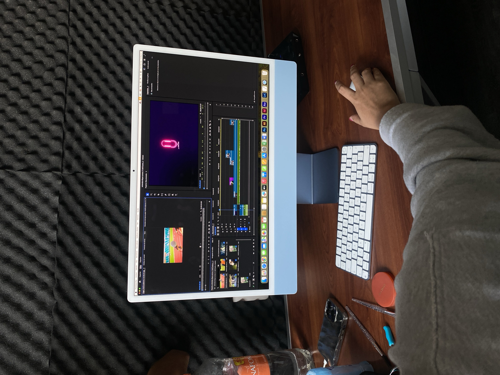 Here's a concise description of the image:
A medium shot, taken from above, shows a person working at a desk. The person's right hand is visible near a white wireless keyboard. They're using an Apple iMac displaying a video editing interface, possibly Adobe Premiere Pro, showing a timeline with video and audio clips. Several icons and a small video preview are on the right side of the screen. Other items on the desk include a cell phone, pens, and a water bottle labeled "NAFON." The desk is brown wood, and the iMac is positioned against a black soundproofing foam panel.