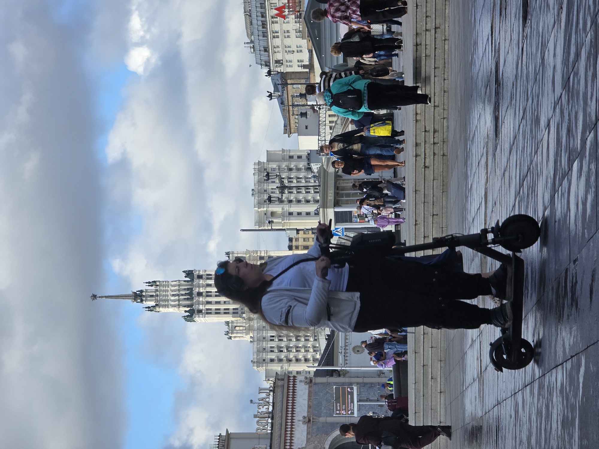 An image captures a street scene in Moscow, Russia, likely near the Komsomolskaya Square, given the presence of the iconic Stalinist skyscraper visible in the background and the architectural style of the buildings. In the foreground, a woman with long, dark hair, wearing a white jacket over a white t-shirt and black pants, rides an electric scooter. She has blue-tinted sunglasses perched on her head. The ground, paved with large square tiles, is wet, reflecting the overcast sky and the surrounding structures.
Behind the woman on the scooter, a group of people are seen walking on a paved area with stairs. The Stalinist skyscraper, a prominent landmark, dominates the background, its ornate spire topped with a star. Several other buildings with classical architecture line the street. The sky is cloudy with patches of blue. Other individuals are visible in the mid-ground, some walking, others seated on steps. A sign with Cyrillic text is also present. The overall atmosphere suggests a bustling urban environment.