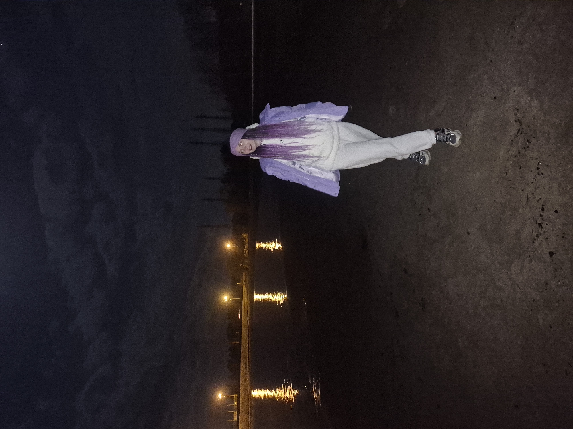 A young woman in a light purple jacket, white pants, and white shoes lies on an asphalt or concrete surface adjacent to a body of water. The woman also wears a light purple hat and her hair appears to be light purple as well. The body of water is lined with lights, and the reflections of those lights are visible in the water. The sky is dark, suggesting it is either night or dusk. Based on the water's edge, the asphalt or concrete surface could be part of a pier, promenade, or walkway alongside the water.