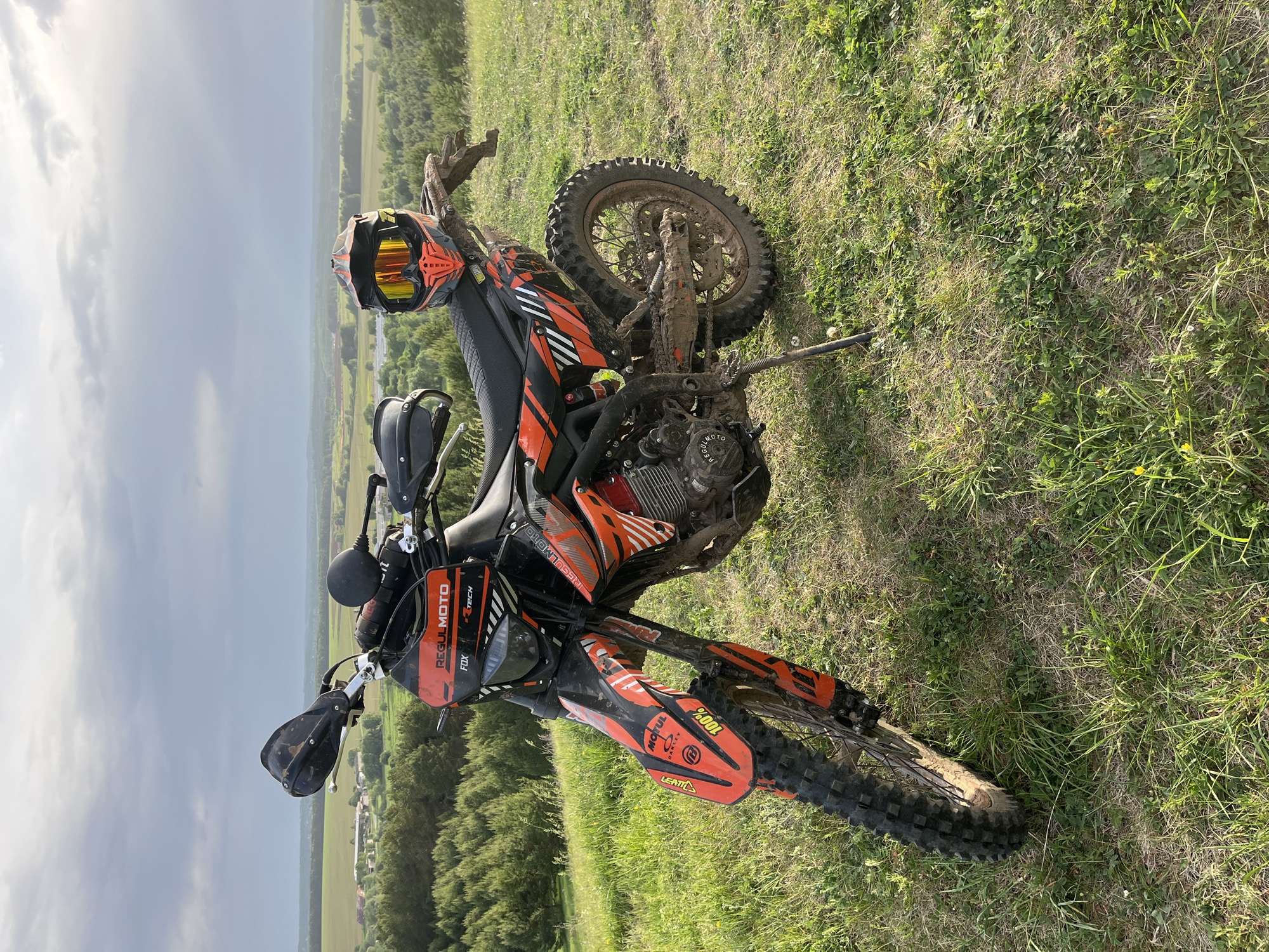 The image shows an orange and black dirt bike lying on its side in a grassy field, possibly a hillside or slope. The bike is covered in mud. The helmet is resting on the handlebars. A stand is visible. The background reveals a landscape of fields, trees, and what appears to be a city or town in the distance. The sky is overcast.