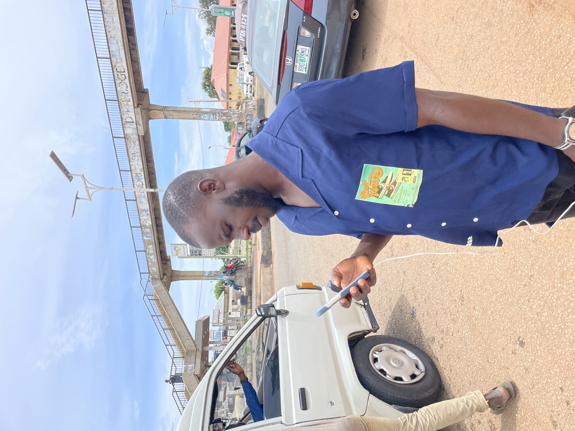 A medium shot captures a Black man in a blue button-down shirt with a graphic print on the left chest pocket, looking down at his phone, which he holds in his right hand. He has a short beard and a light-colored wire is connected to his phone. The man is standing on a textured, reddish-brown pavement, next to the open passenger door of a white van. A portion of the van's wheel is visible. In the background, an elevated pedestrian bridge with railings spans across the scene. Graffiti is visible on the side of the bridge. A traffic light pole stands near the bridge, and a dark-colored sedan with a license plate reading "RBC 879MQ" is parked to the right of the man. Other vehicles and buildings are faintly visible further in the background, suggesting an urban environment. The sky above is partly cloudy. The scene appears to be set in Gura Top, Nigeria.