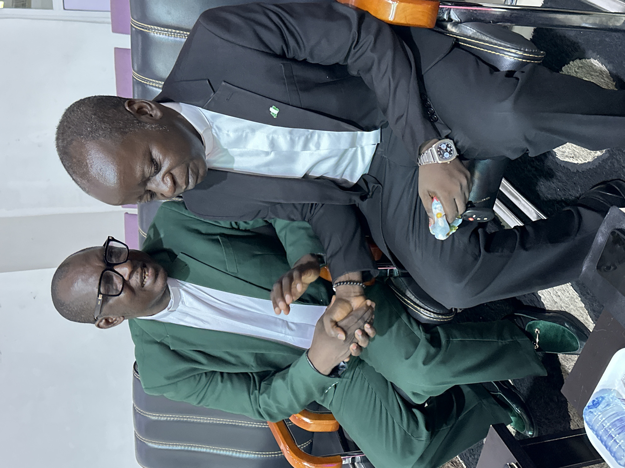 Two men are seated in black leather chairs, shaking hands. The man on the left wears a green suit, a white collar, and black-rimmed glasses. The man on the right wears a black suit, a white collar, and a Nigerian flag pin on his lapel. The setting appears to be an indoor waiting area, possibly in Goni Gara, Nigeria, suggested by the mention of the location, though the image itself does not provide explicit visual cues for this specific locale. The lighting is artificial, and the background is a muted wall with some purple seating visible in the distance.