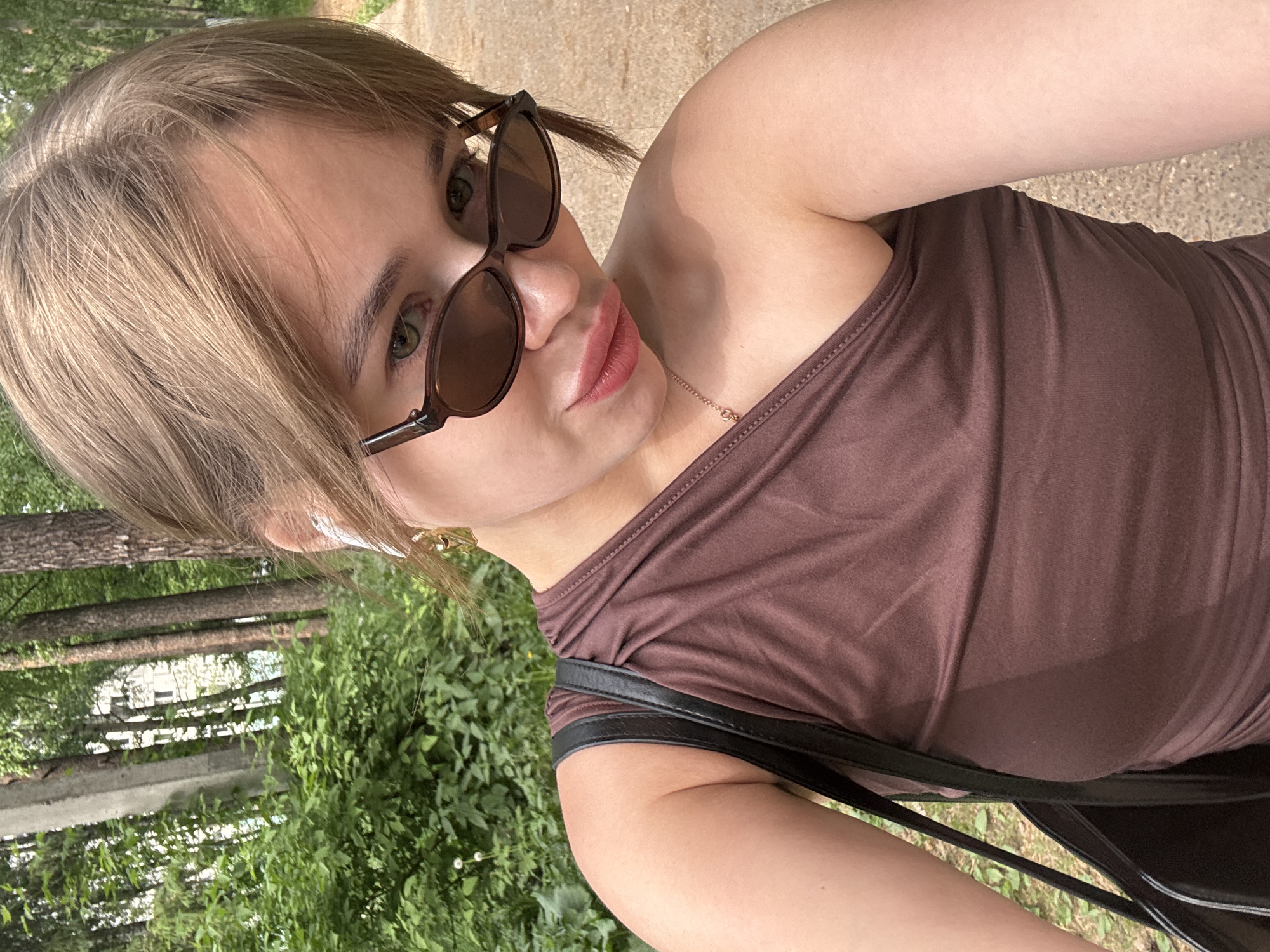 A woman is taking a selfie outdoors. She has short blonde hair, is wearing sunglasses and a brown one-shoulder top, and has a black bag slung over her shoulder. The background consists of trees, foliage, and a paved path, suggesting a park or green space. Based on the query parameters, the location is the city of Chaikovsky, Russia.