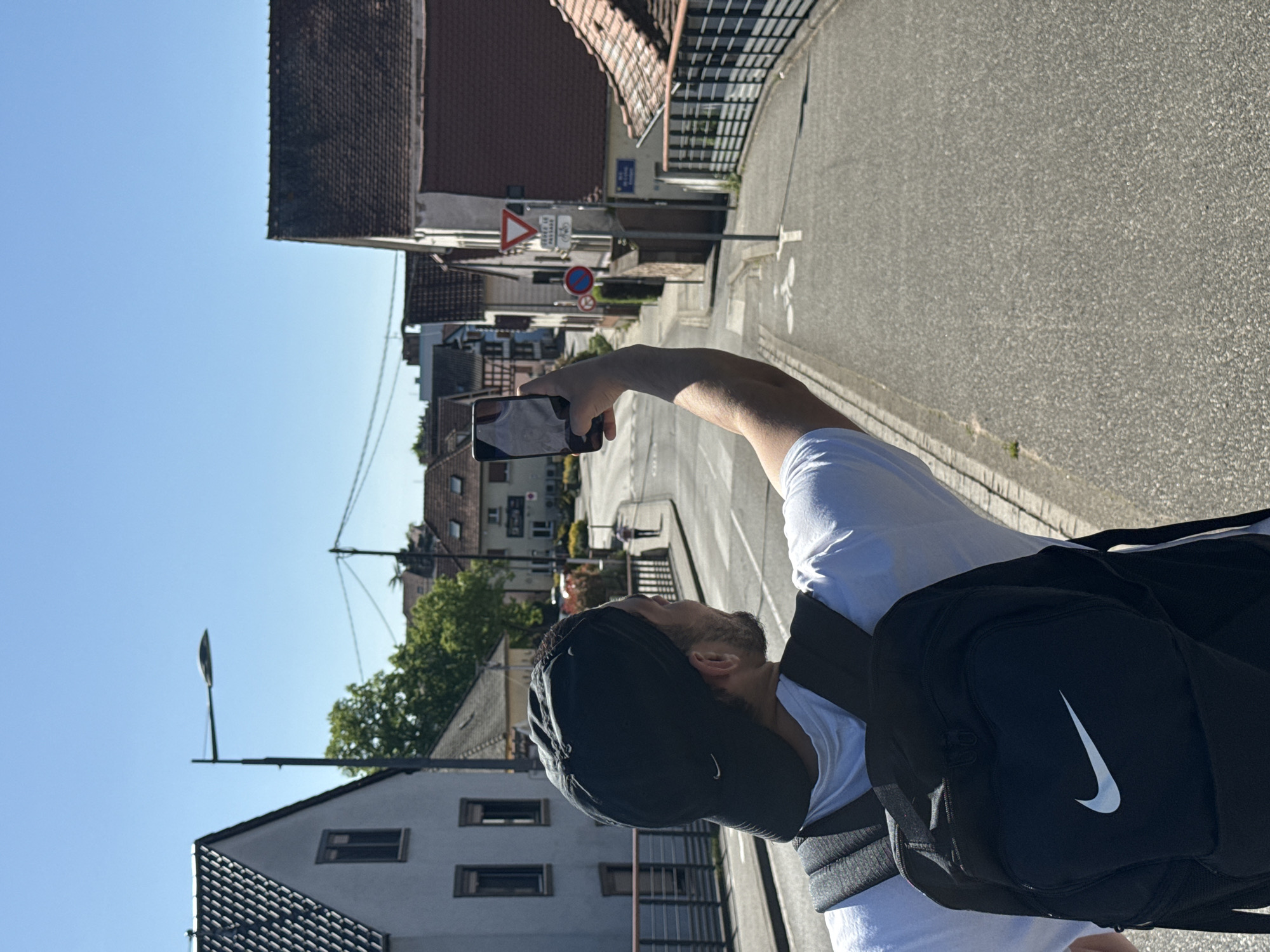 A man is facing away from the camera and holding up his cell phone to take a photograph in Hoenheim, France. The man is wearing a white t-shirt, a black baseball cap, and a black Nike backpack. The street is lined with buildings, including houses and apartments. There is a street sign indicating a "yield" intersection. The street is paved with asphalt. The sky is blue.