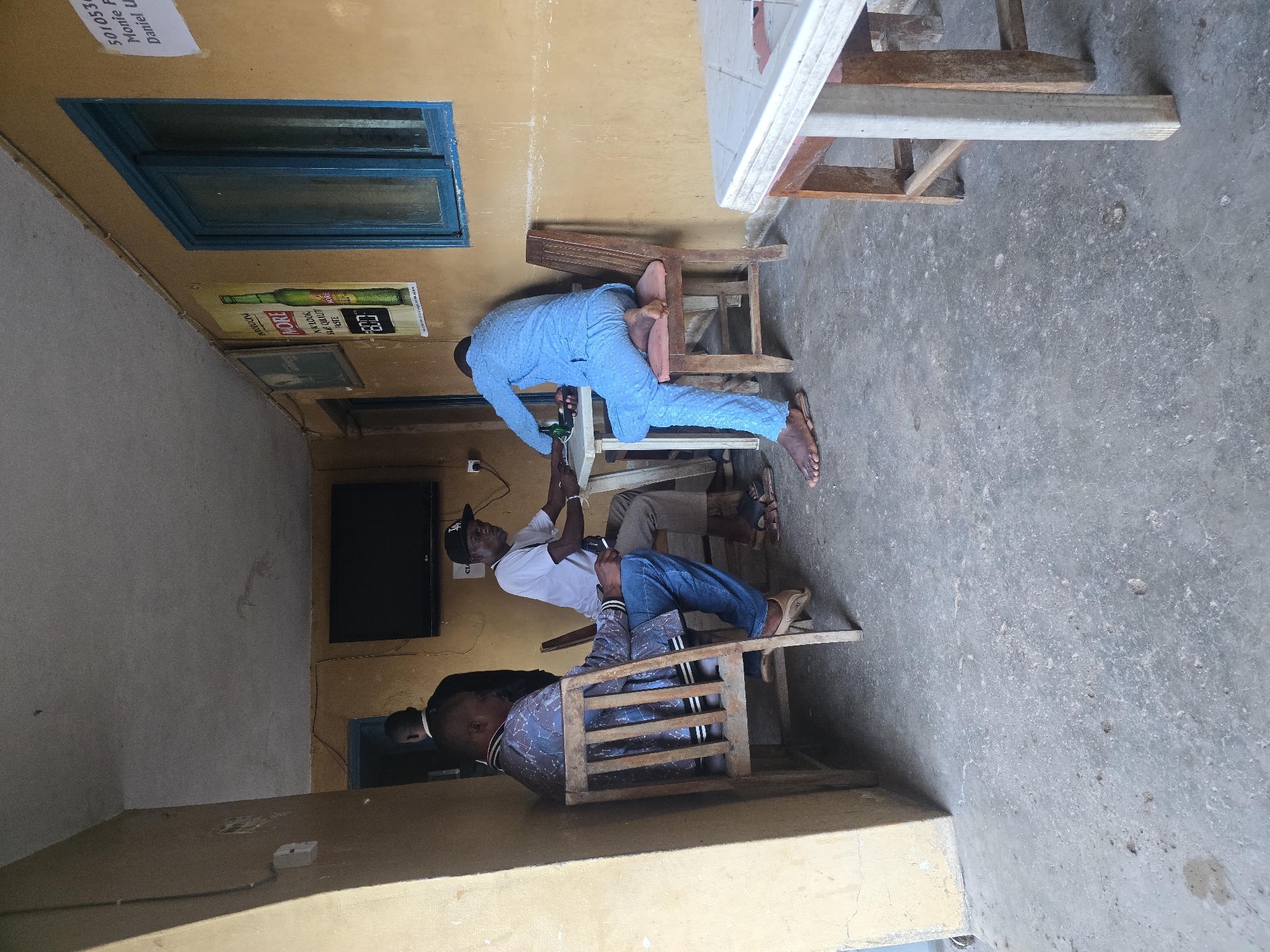 In a partially open-air establishment, possibly a bar or eatery, located in the city of Jos, Nigeria, four men are observed. Two of the men are seated at a white plastic table, engaged in conversation and consuming beverages. One man, wearing a white t-shirt and a black baseball cap, is looking down at the table. The second man, dressed in a light blue, textured tunic and trousers, is reaching towards a green bottle on the table. Another man, wearing a patterned jacket and blue jeans, is seated in a wooden chair facing away from the camera. A fourth man, seen from the back, is standing near a doorway in the background. The environment appears worn, with a concrete floor, yellowish walls, and a visible television mounted on the wall. Advertising posters are displayed on the wall, and a blue-framed window is present. The overall impression is one of casual social gathering in a modest setting.
