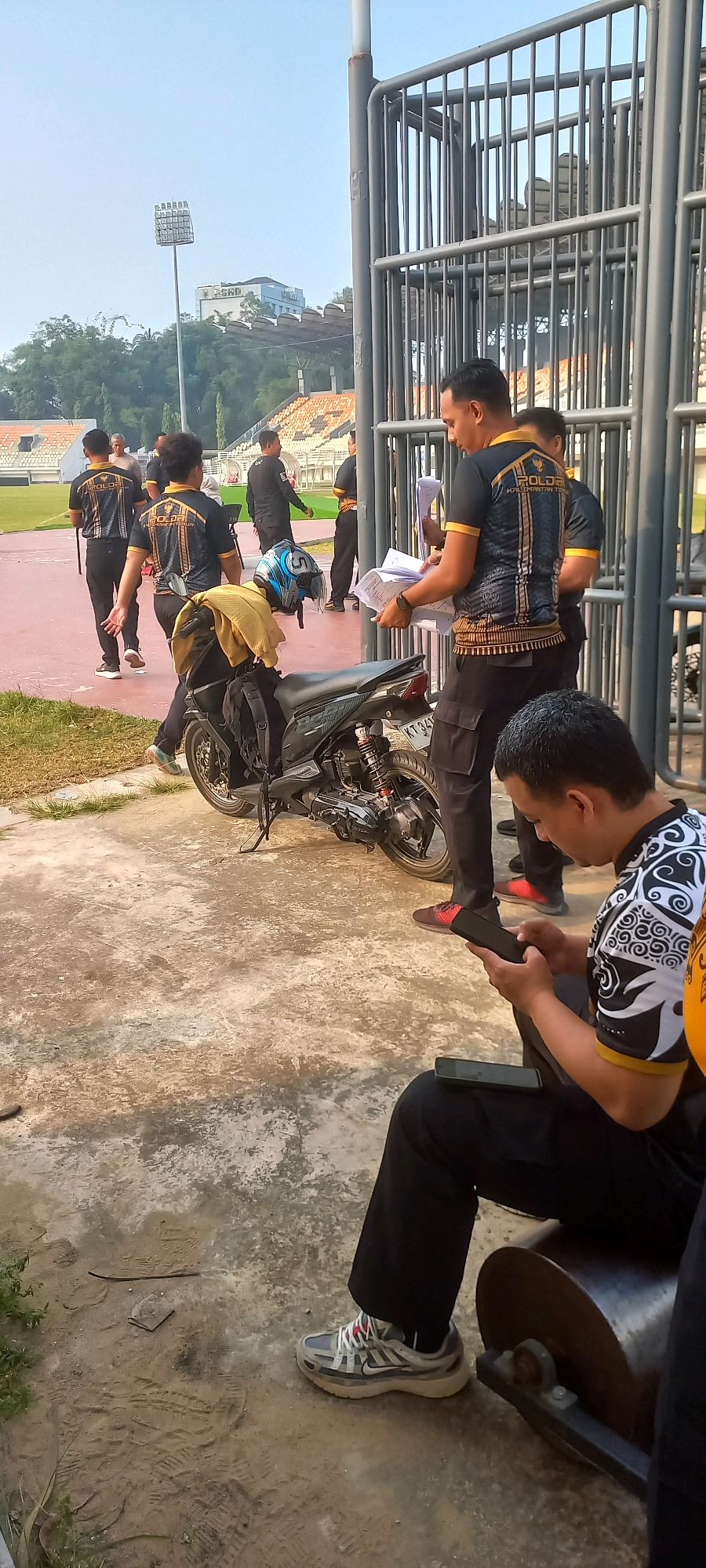 The image depicts a scene at the Gelora Kadrie Oening stadium in Samarinda, Indonesia, on a clear day. In the foreground, a person is seated and looking at their smartphone, with another phone on their lap. To their left, a black motorcycle is parked. Behind the motorcycle, a group of men in matching black and gold uniforms are standing near a metal fence. Some of them are holding papers, suggesting an official capacity. The stadium's seating area and a light tower are visible in the background, with trees and buildings on a hill beyond. The ground is a mix of concrete and dirt. The men in uniforms appear to be security personnel or staff associated with the stadium.