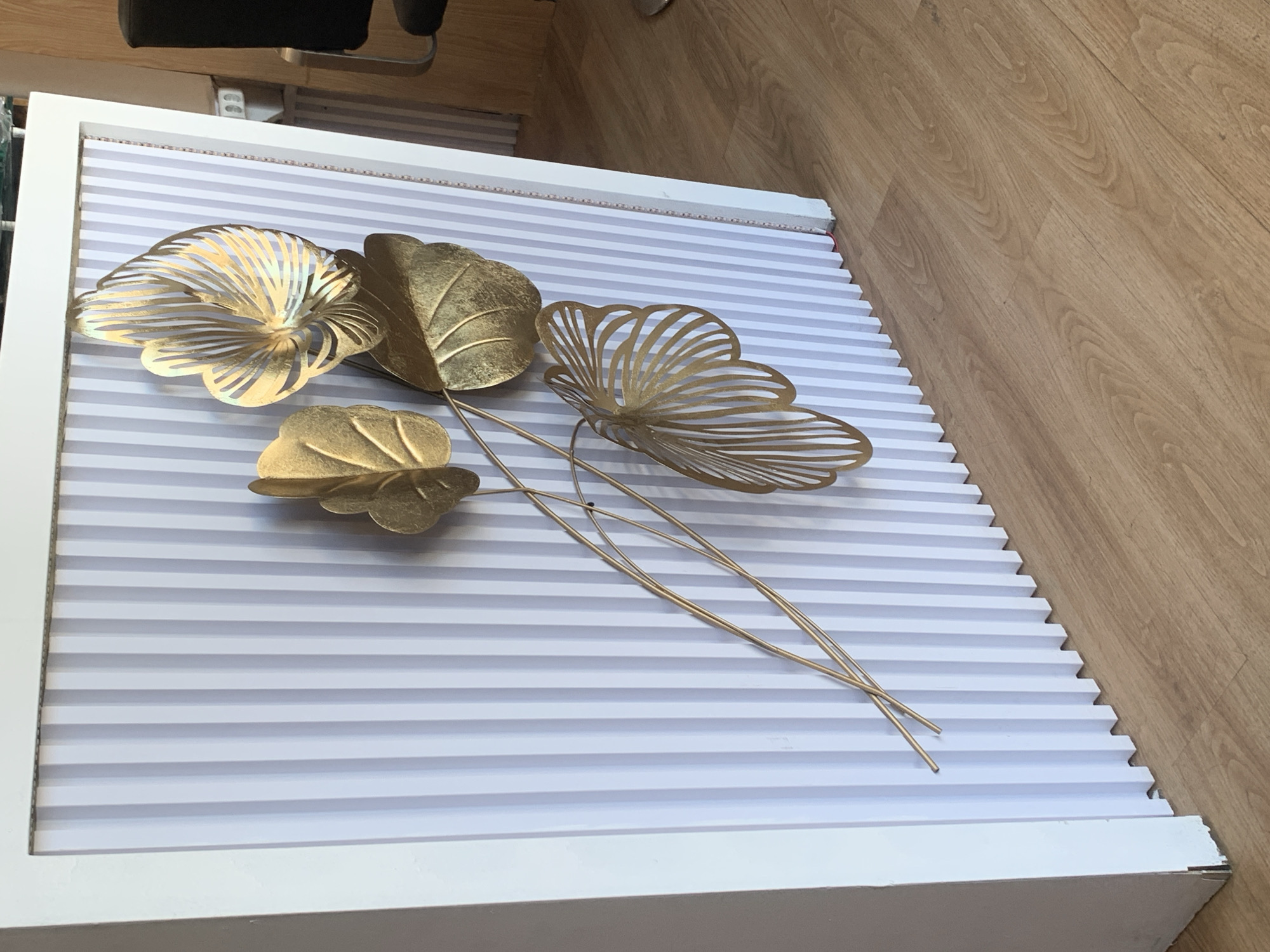 A close-up, eye-level shot showcases a portion of a white frame enclosing a fluted wall panel with alternating light gray and white vertical stripes. A gold metal art piece, resembling a cluster of leaves and branches, is attached to the panel. Four distinct leaf-like shapes are visible: two solid, textured gold leaves, one larger and curved, the other smaller and ovular, positioned above and to the left of the larger one. A third gold leaf, also solid and textured, is larger and more prominently displayed, its broad, curved shape dominating the center-right of the image. Below this, a fourth, larger gold element is intricately cut with delicate, radiating lines, resembling a feather or a stylized fan. Thin gold wire branches connect these elements, extending diagonally from the lower left to the upper right. The bottom edge of the fluted panel meets a light brown wooden floor, the planks laid horizontally. To the right of the panel, a sliver of a dark chair is visible, and behind it, a white wall with electrical outlets and a wooden door frame. The lighting is moderate, casting soft shadows that highlight the metallic sheen of the art piece and the texture of the wooden floor. The scene appears to be an indoor setting, possibly a commercial space or a decorative display, in Denpasar, Indonesia.