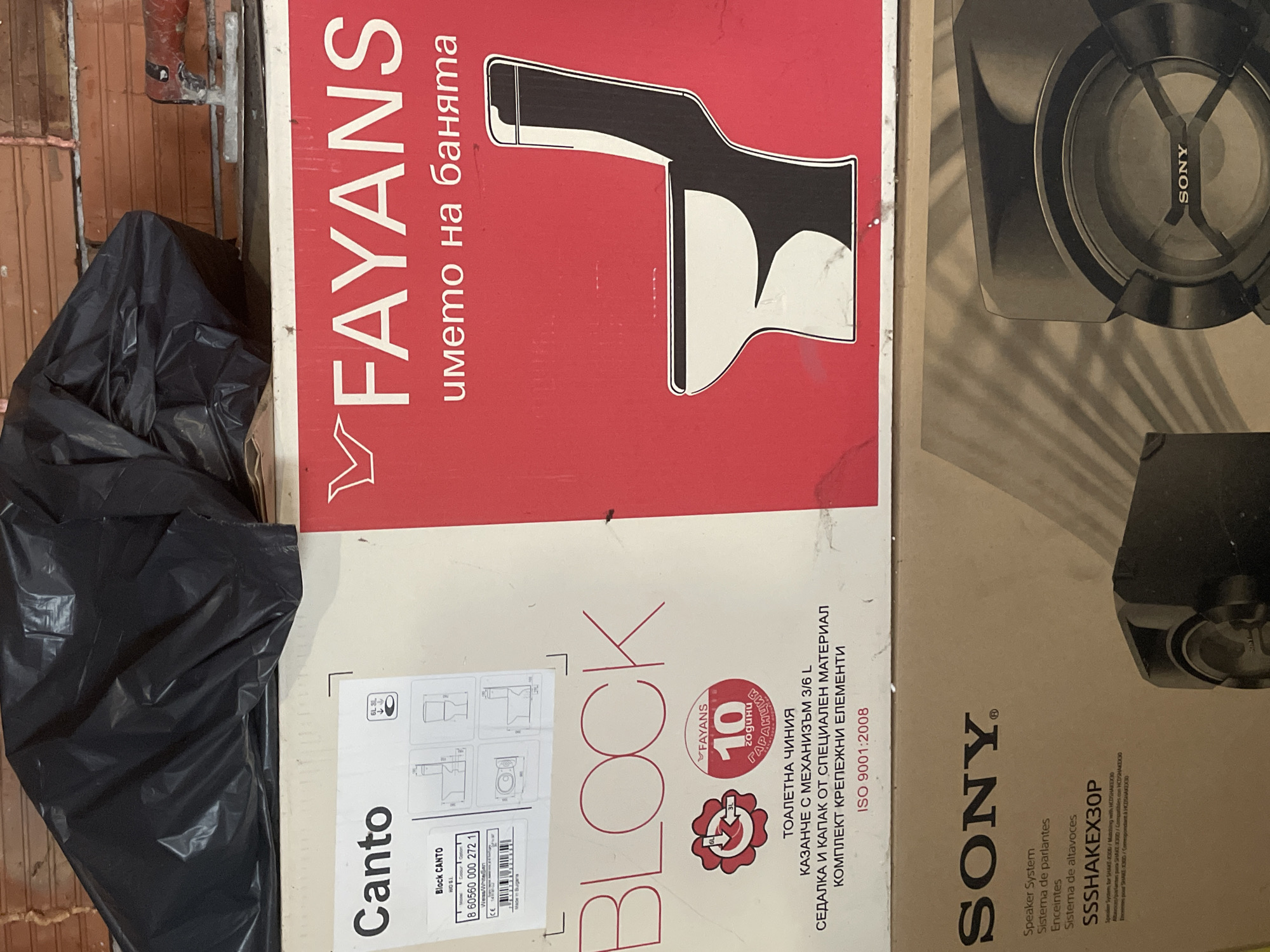 The image shows two stacked cardboard boxes and a black plastic bag in what appears to be an indoor setting, possibly a storage area or a construction site in Ihtiman, Bulgaria.
The top box is predominantly red and white, branded "FAYANS" with the slogan "името на банята" (the name of the bathroom). It features a stylized illustration of a toilet and is identified as "BLOCK CANTO". The text indicates it contains a "ТОАЛЕТНА ЧИНИЯ" (toilet bowl), a "КАЗАНЧЕ С МЕХАНИЗЪМ 3/6 L" (cistern with 3/6 L mechanism), a "СЕДАЛКА И КАПАК ОТ СПЕЦИАЛЕН МАТЕРИАЛ" (seat and lid made of special material), and a "КОМПЛЕКТ КРЕПЕЖНИ ЕЛЕМЕНТИ" (set of fastening elements). It also prominently displays "10 години ГАРАНЦИЯ" (10 years warranty), "ISO 9001:2008", and "Made in Bulgaria". A barcode and product number "8 60560 000 272 1" with "Weiss/White/Br" for color are visible.
Beneath and to the right of the red box is a brown cardboard box, featuring the "SONY" logo. This box is for a "Speaker System" with the model "SSSHAKEX30P". Additional text in Spanish and French, "Sistema de parlantes" and "Enceintes", confirms its contents as speakers. A portion of the speaker's grille is visible.
To the left, a crumpled black plastic bag covers an unseen object. In the background, a brick-like wall structure is partially visible, hinting at a raw or unfinished environment. No people are present, and no specific activity is occurring; the items seem to be stored.