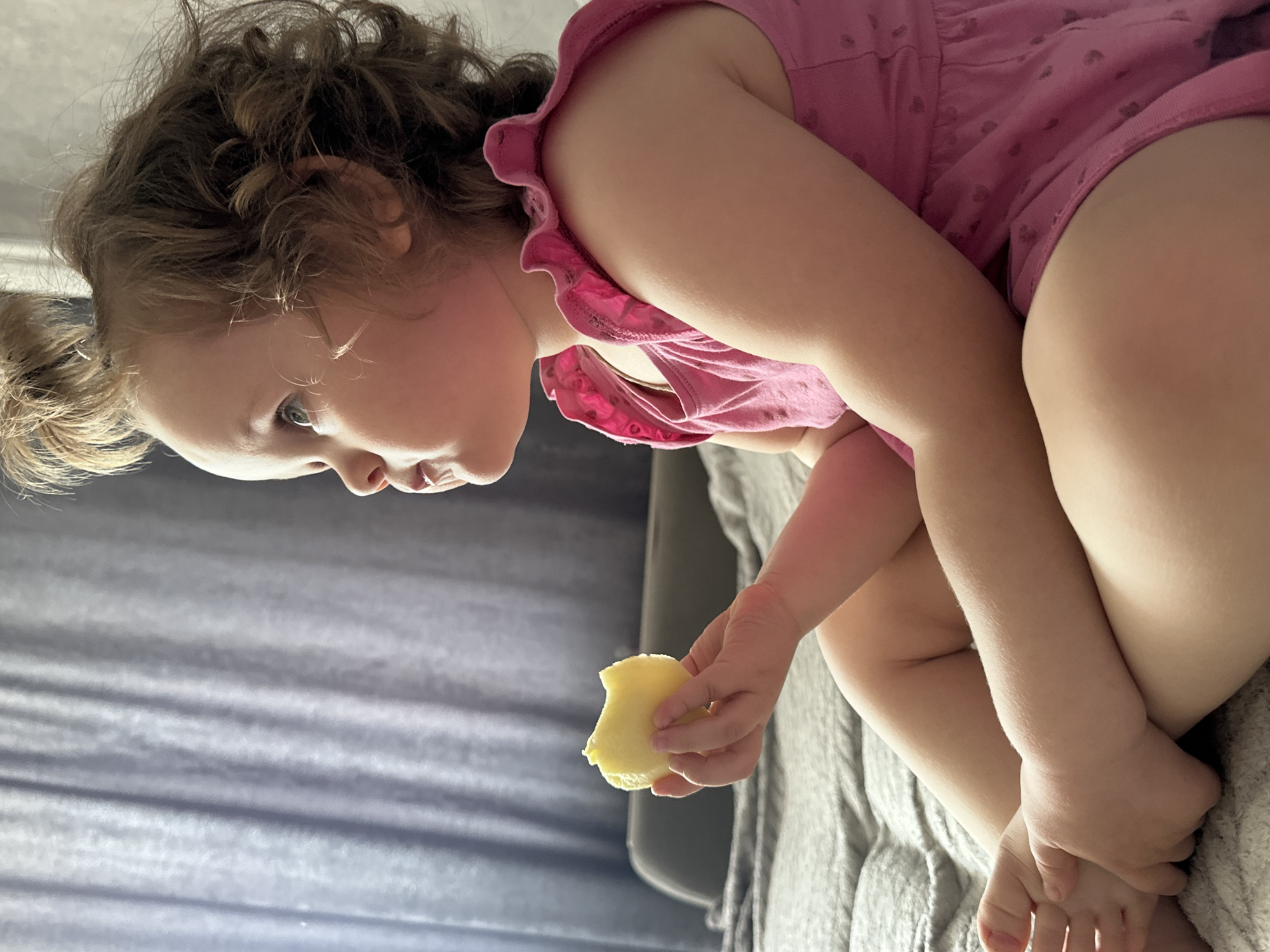 A young child with curly hair is seated, holding a piece of yellow fruit. The child wears a pink dress with a ruffled neckline. The background includes a textured gray surface and a hint of a window with light filtering through. There is no information available to indicate that the location is the city of Chaikovsky, Russia.