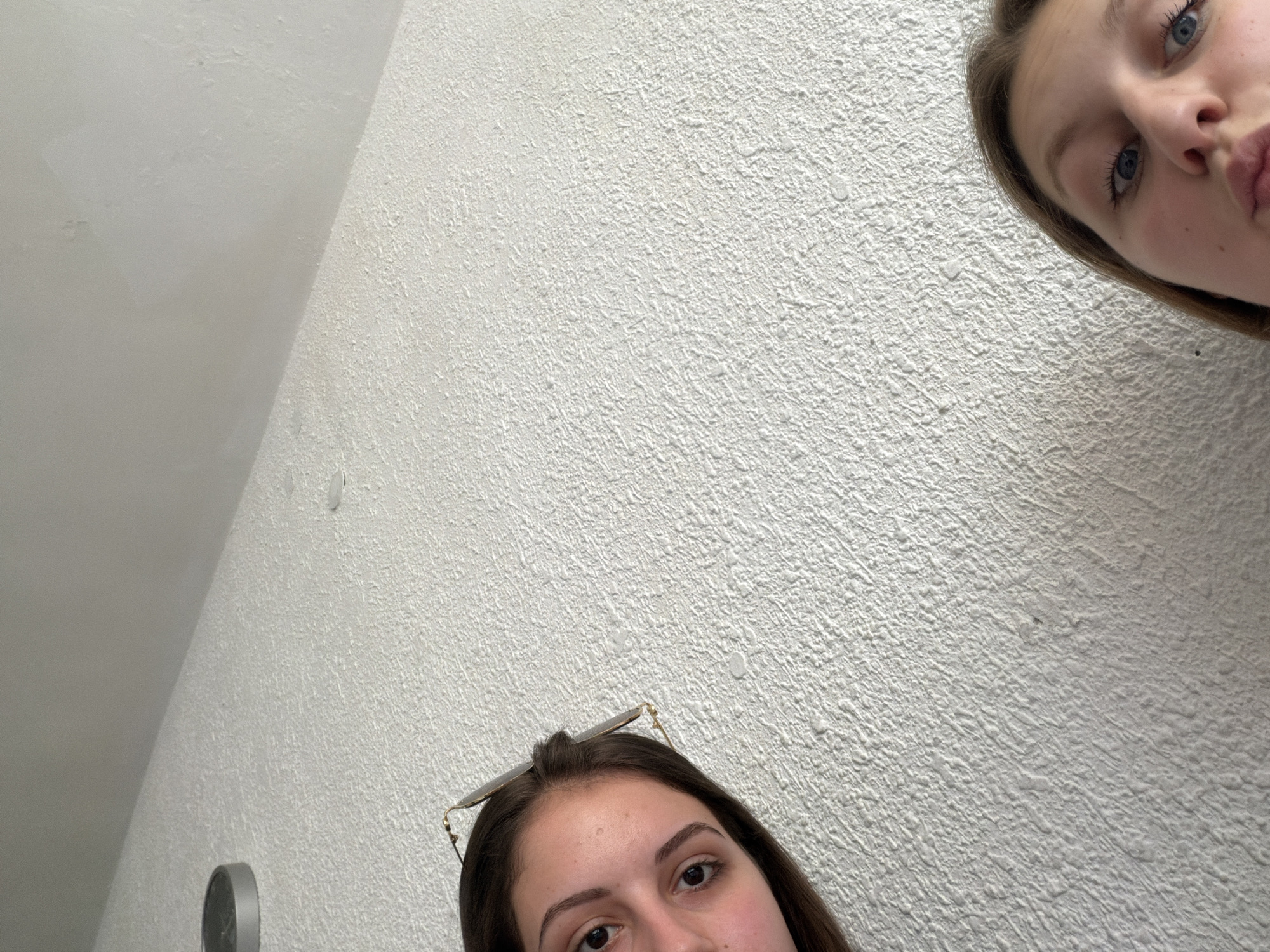 Two young females are captured in a low-angle, selfie-style photograph. The background consists of a textured, off-white wall that extends upwards, meeting a lighter ceiling. On the upper left portion of the wall, a round, metallic object is visible, possibly a smoke detector or a light fixture. The first female is on the left side of the frame, with her head angled towards the camera. She has dark hair and is wearing sunglasses perched on her head. Her eyes are open, and she appears to be looking directly at the camera. The second female is positioned in the lower right corner of the image, with her head tilted forward. She has blonde hair and her eyes are wide open, with her mouth slightly pursed as if puckering. The image does not provide enough information to determine the specific location in Burgas, Bulgaria.