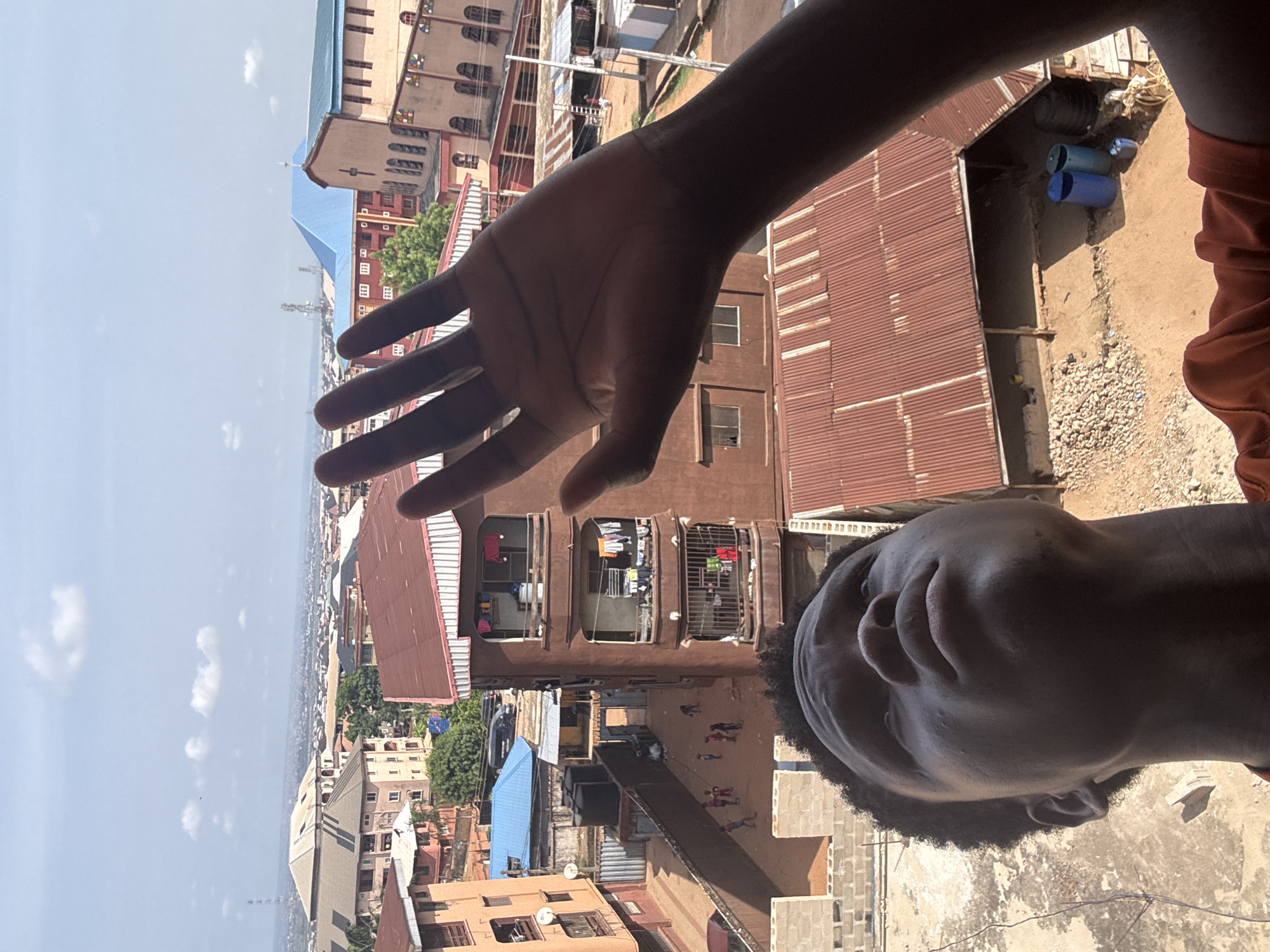This image, rotated 90 degrees clockwise, presents a high-angle view of a densely populated urban landscape under a bright, clear sky. In the foreground, a dark-skinned person's face is visible on the right, looking upwards towards the camera with a neutral expression. Their outstretched left hand, palm facing outwards, occupies the central left portion of the frame, partially obscuring the view. A hint of an orange/brown garment is visible on the person's right shoulder.
The background reveals a bustling city scene, consistent with the context of Onitsha, Nigeria. Numerous buildings with reddish-brown corrugated iron roofs are prominent. A multi-story brown residential building directly below the hand features windows with metal grates and clothes hanging on balconies. Further into the mid-ground, a larger building with a distinct light blue roof and facade, characterized by arch-shaped windows and some colorful accents, possibly a church or public building, is visible.
Below the buildings, a dusty ground area shows several children walking or playing, alongside some blue barrels or containers. The distant horizon is filled with a sprawling cityscape, and communication towers or masts can be seen. The bright sunlight and strong shadows indicate it is daytime with good weather. There is no visible text in the image.