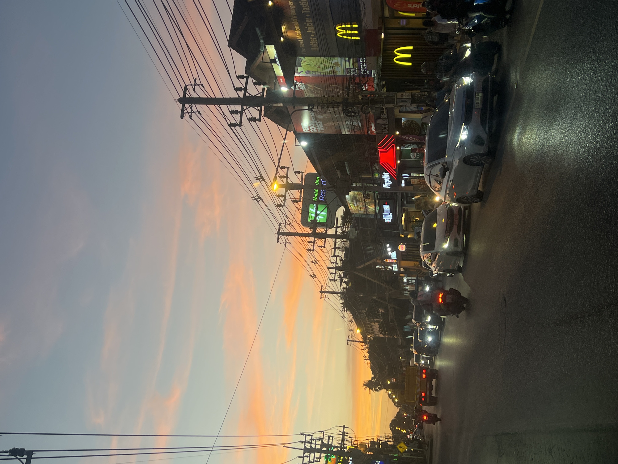 The image depicts a street scene in the city of Ko Samui District, Thailand, during sunset. The sky is painted with hues of orange and pink, with wispy clouds. A busy street is lined with buildings, some featuring illuminated signs for fast-food restaurants like McDonald's and Pizza Hut, and a Holiday Inn. Overhead, a complex network of power lines crisscrosses the sky, supported by utility poles. Traffic is moderate, with cars and motorcycles on the road. A yellow truck is visible in the distance. The overall atmosphere suggests a vibrant urban environment at dusk.