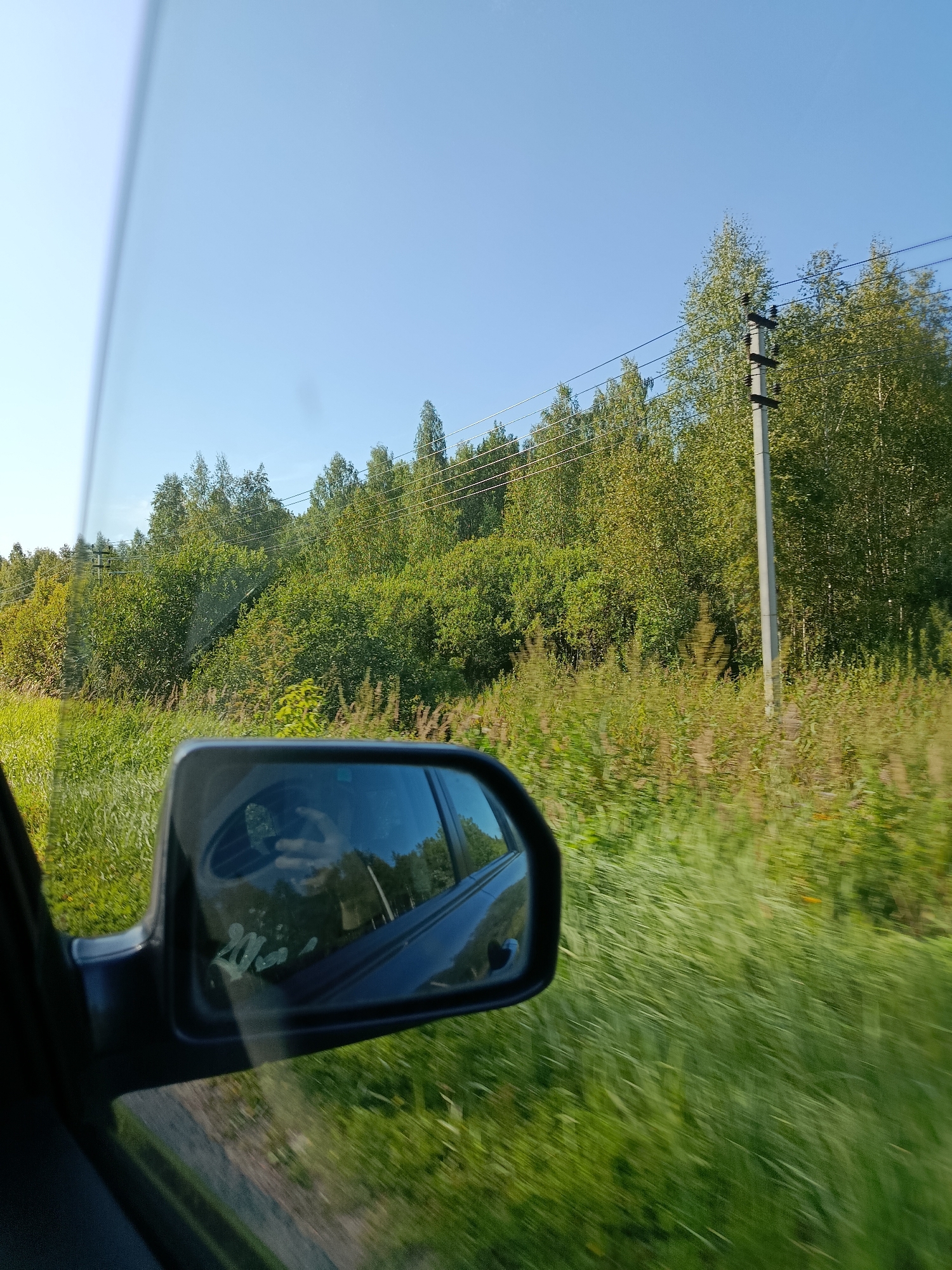 The image captures a view from inside a vehicle, looking out the side window and into the side-view mirror. The scene is set during the day under a clear blue sky, suggesting a sunny environment. The landscape outside consists of lush green vegetation, with a dense forest of trees forming the background. Power lines are visible stretching across the scene, supported by a utility pole. The side-view mirror reflects the interior of the car, including a person's hand holding a phone. Motion blur is evident throughout the image, indicating that the vehicle is in transit, likely on a road. The overall impression is a snapshot of a journey through a natural, rural setting, possibly on the outskirts of Верея, Russia, given the context.