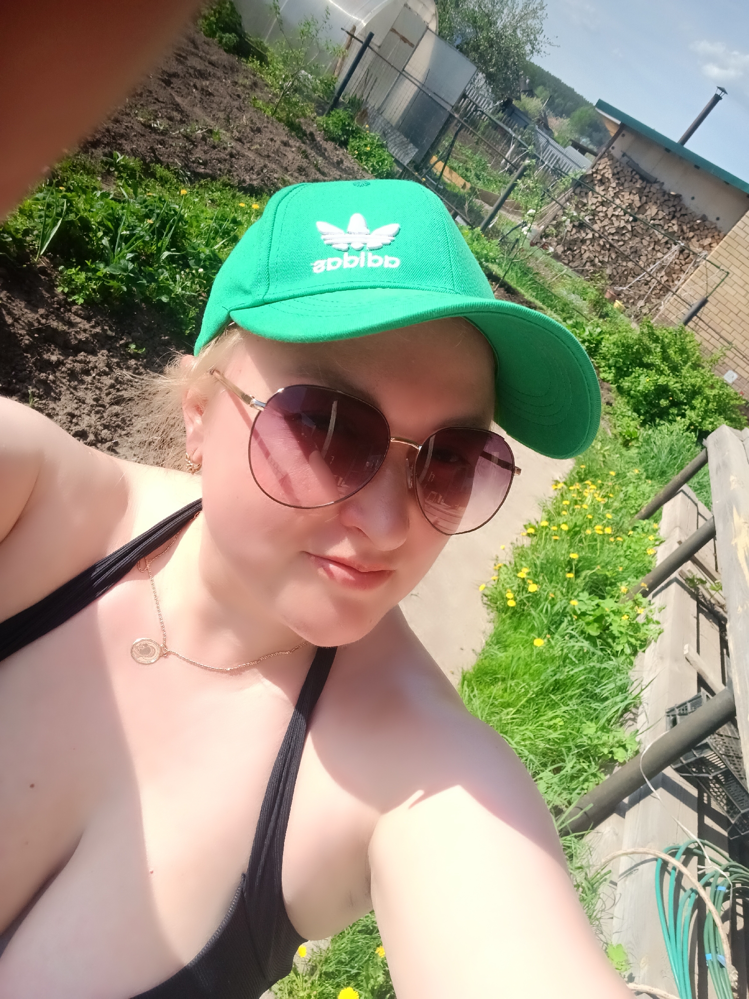 A selfie of a person wearing a green "Adidas" cap and brown sunglasses, positioned at an angle. The person is wearing a black top and a gold necklace. The background includes a garden with dandelions, a building made of brick, and a pile of firewood. The city of Chaikovsky, Russia, is not identifiable in this image.