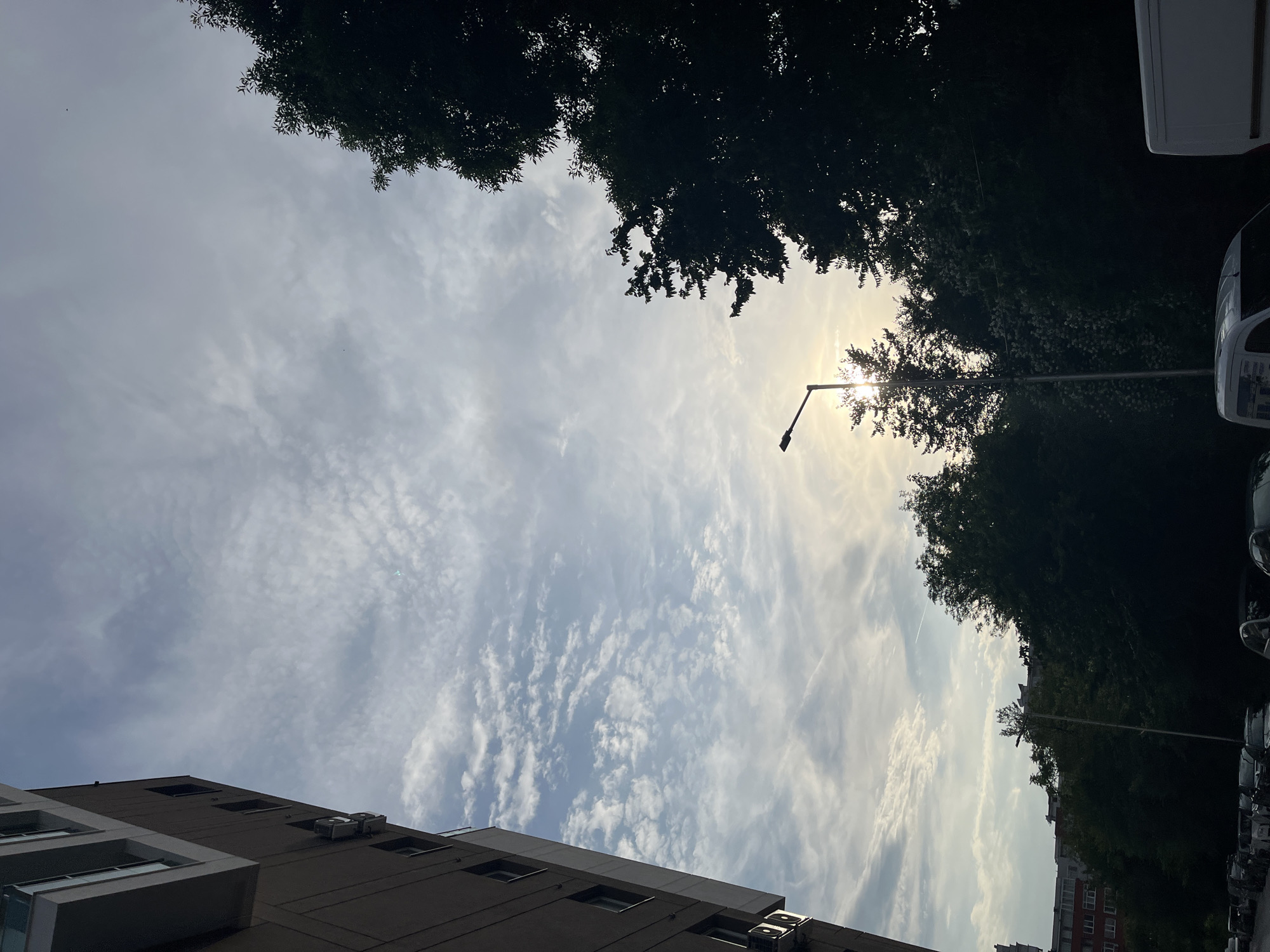 The image presents an upward-looking view of a partially cloudy sky, dominated by white and light grey clouds with interspersed patches of blue. The sun, a bright glare, is positioned in the upper right quadrant, partially obscured by the dark foliage of trees. A dark street lamp post, silhouetted against the bright sky and sun, extends an arm into the frame.
On the left side, the upper stories of a modern, multi-story building are visible, featuring a dark brown or grey facade with multiple rectangular windows and white-framed sections. Several air conditioning units are mounted on its exterior. On the right, the tops of dark green trees form a dense canopy along the edge, and the upper parts of parked vehicles, including a white van and other cars, are discernible. A faint white contrail is visible against the blue sky in the upper left. The scene captures an urban or suburban environment in Veliko Tarnovo, Bulgaria, on a clear to partly cloudy day. The time appears to be either morning or late afternoon, given the sun's angle and intensity. No specific activity or event is taking place, nor is any text visible.