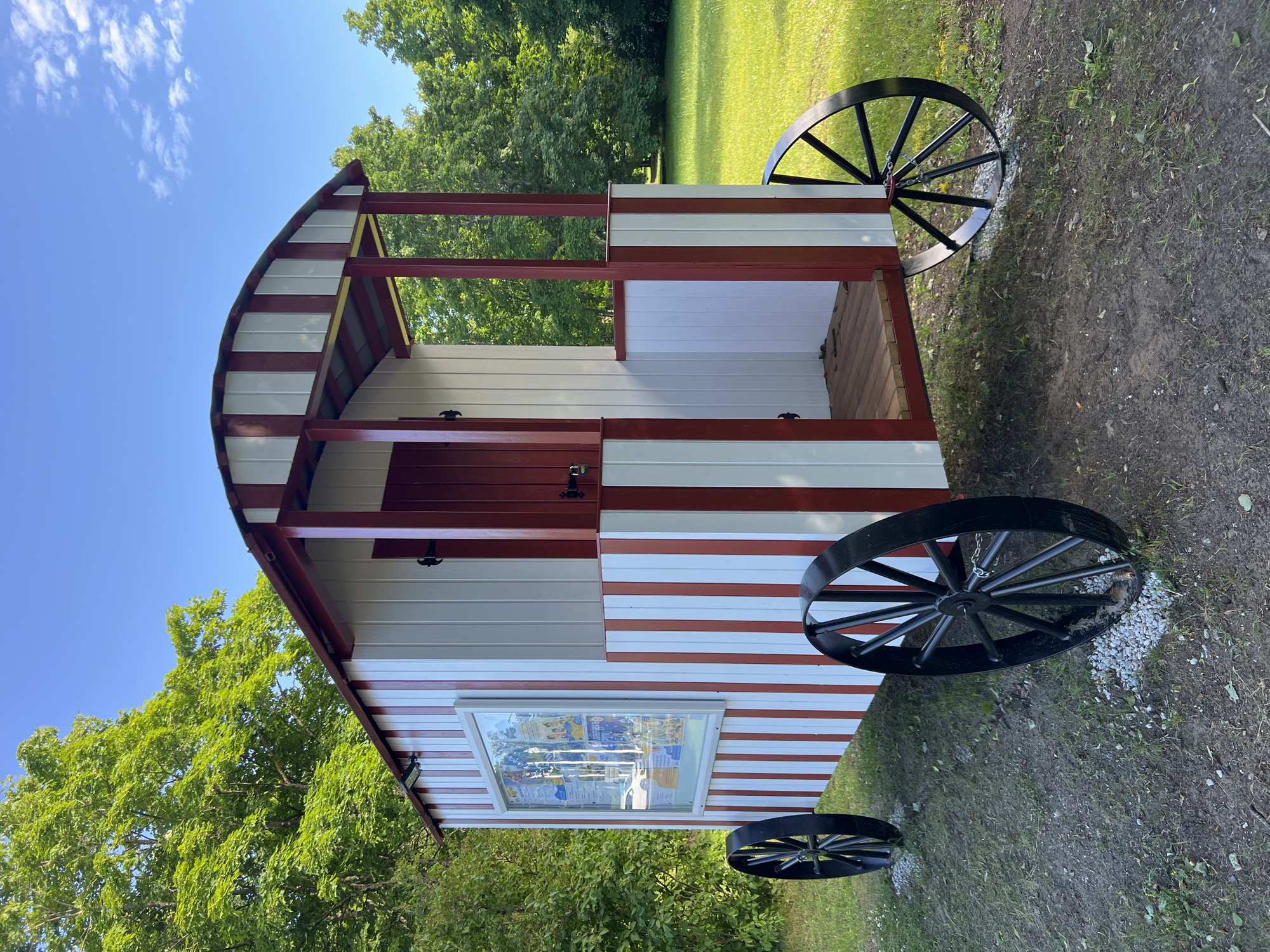 A small, red and white, wagon-like structure sits on a grassy and dirt area. The structure has a rounded, arched roof and is supported by red beams. It features two large black spoked wheels on each side, attached to the structure by chains. A window on the side of the wagon displays what appears to be a poster or information board with multiple images and text, though the text is illegible. The wagon is painted with alternating red and white horizontal stripes on its lower half. The setting is outdoors, with lush green trees and a clear blue sky visible in the background, suggesting a sunny day. The ground is a mix of grass and exposed dirt, with some scattered white gravel near the wheels. The overall scene appears to be a park or a historical site. No people are visible in the image. The time of day appears to be daytime. The location context is Narva-Jõesuu, Estonia.