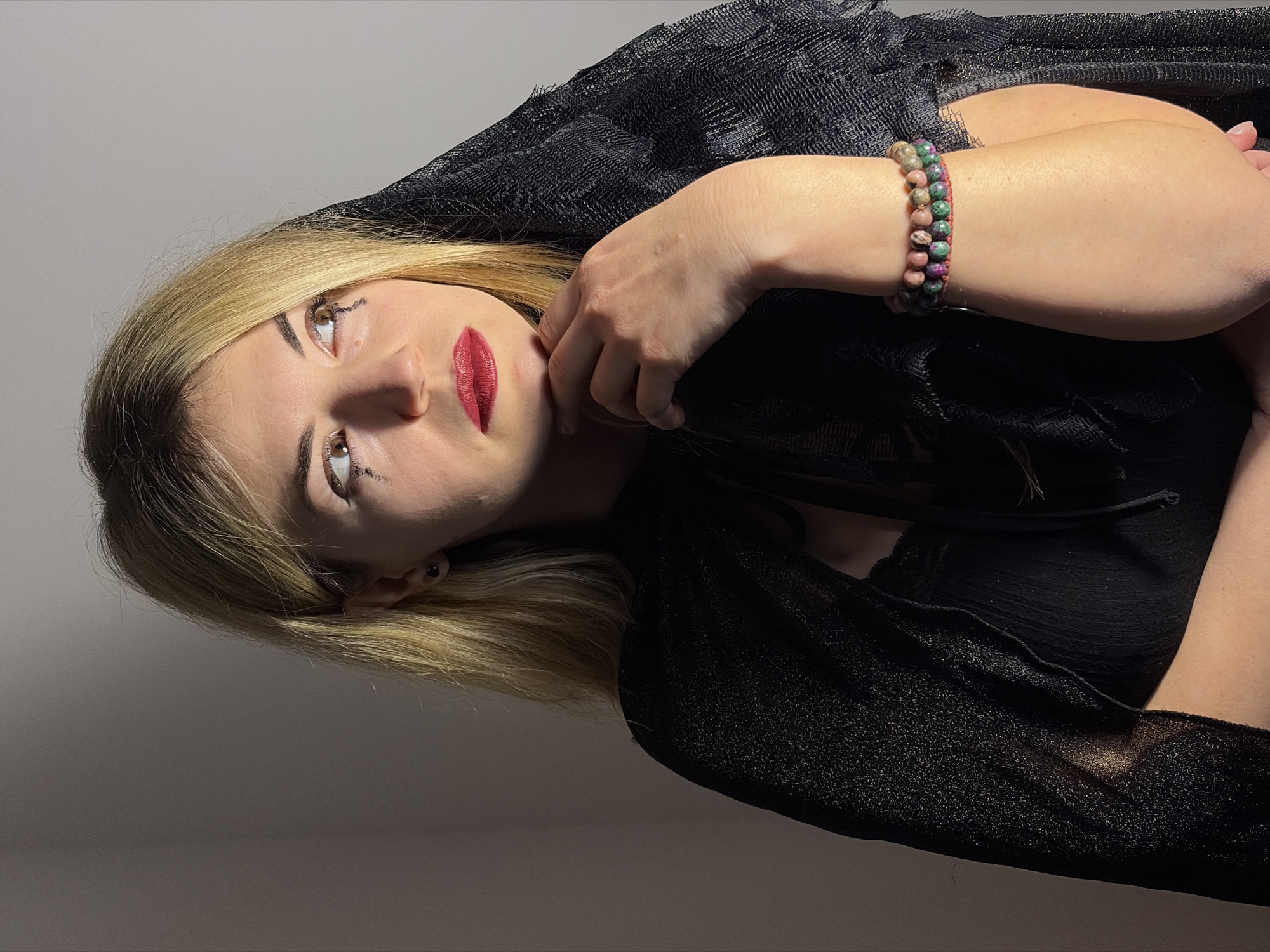 A woman with blond hair is pictured in a dimly lit environment, presumably indoors, as suggested by the plain background. The woman is wearing red lipstick and has dark smudges beneath her eyes. She is also wearing a black, lace-like garment that drapes around her head and shoulders. A beaded bracelet adorns her wrist. The scene appears to be photographed in a dimly lit place, perhaps a studio. Location cannot be ascertained as Budva, Montenegro.