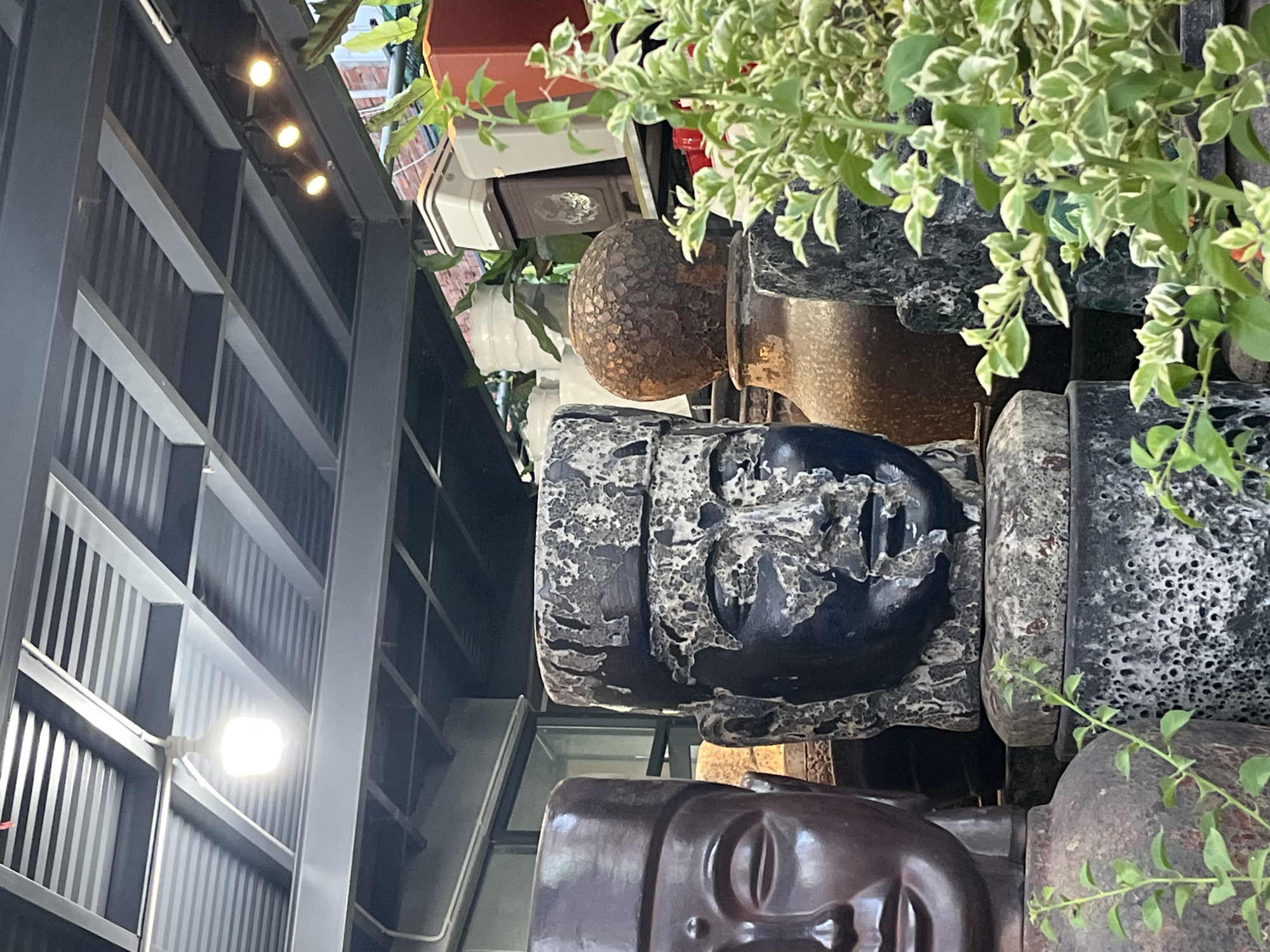 This image depicts an outdoor display of various large decorative objects, likely in a garden center or a similar retail environment, in Bac Nha Trang Ward, Vietnam. The foreground is dominated by several large busts and vases. On the left is a dark, smooth bust with closed eyes and a serene expression. To its right is a rough, dark-colored bust with a textured, weathered surface, featuring a human face. Behind these busts, other objects are visible, including a spherical, textured vase and more textured, dark-colored items that appear to be pedestals or bases. In the mid-ground, there are several planters and decorative pots, some of which are white and stacked. A leafy green plant with variegated leaves is prominently featured on the right side of the frame, obscuring some of the background elements. The background reveals a corrugated metal roof structure supported by metal beams, with lighting fixtures attached to it, suggesting an outdoor covered area. The overall scene is a collection of decorative garden items.
