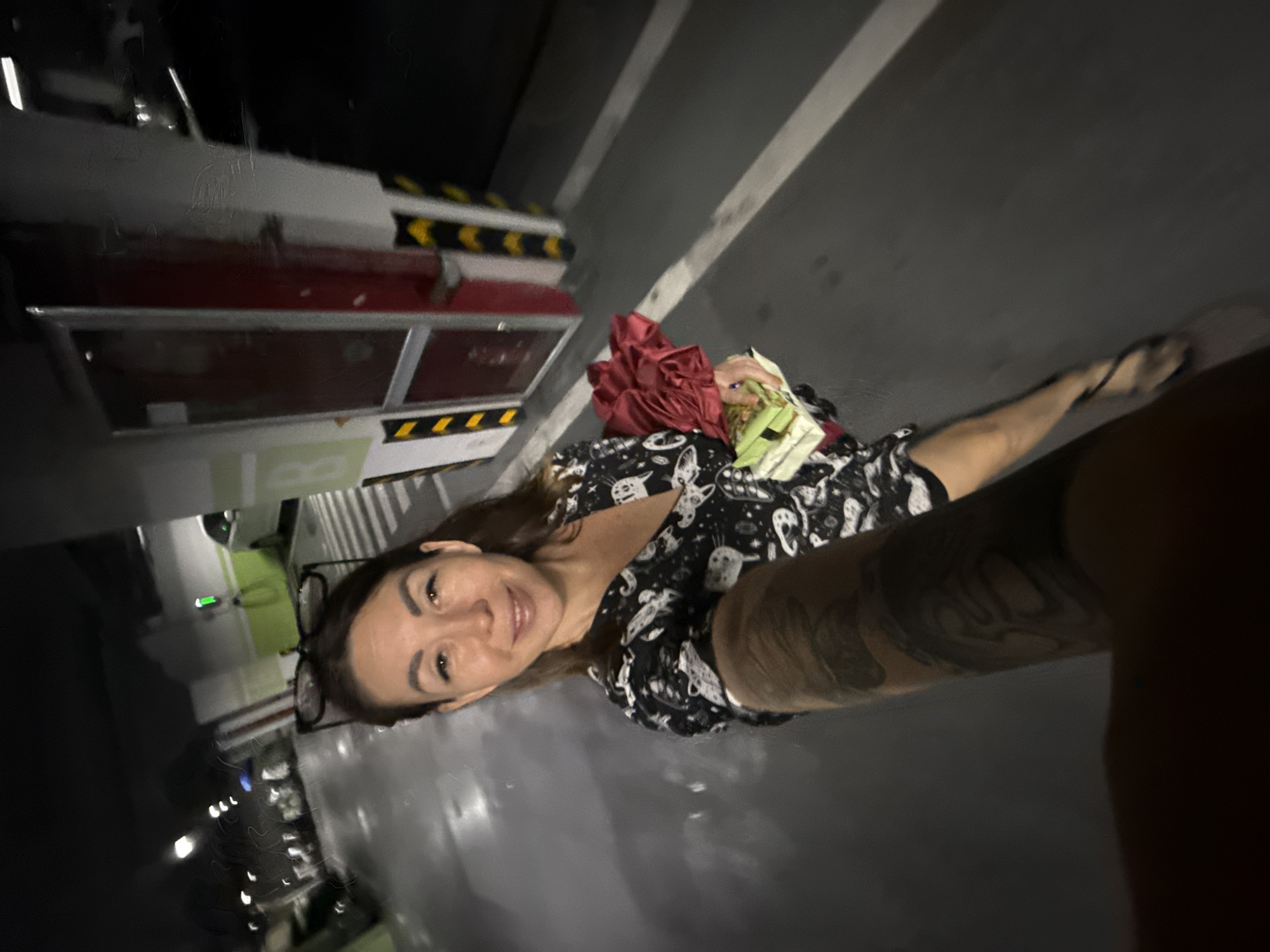 In a dimly lit indoor parking garage, a woman with brown hair and glasses perched on her head smiles at the camera as she holds a bouquet of red flowers and a pack of green items. She is wearing a black and white patterned dress and is holding a smartphone to take a selfie. Her left arm, covered in tattoos, is extended forward. The parking garage appears to be in the city of Huizhou, China, as indicated by the car charging station labeled with green and white markings and the overall infrastructure consistent with the region. The floor is marked with white lines, and the walls are painted white and grey, with red and yellow safety markings on columns. Several vehicles and the entrance to an elevator or stairwell are visible in the background.