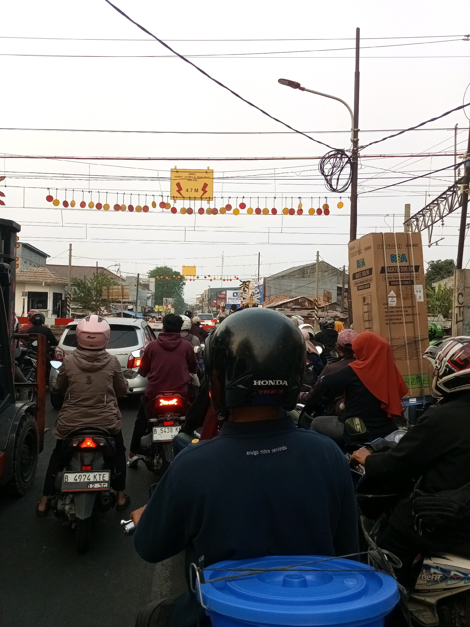 The image depicts a congested street scene in Bekasi, Indonesia, characterized by heavy motorcycle traffic. In the foreground, three individuals on motorcycles are visible from behind. The rider in the center wears a pink helmet and a brown jacket, their motorcycle's license plate reads "B 4974 KTE". To their right, another rider in a maroon hoodie is seated on a motorcycle with a visible license plate "B 5438 KK". The motorcycle furthest to the right has a rider wearing a black helmet. In the background, numerous other motorcycles and a silver car are observed, indicating a traffic jam. Buildings line the sides of the street, with overhead power lines and decorative garlands suspended across the road. A large cardboard box with the text "RSA AGATE 300.IN" is prominently displayed on the right side of the frame, suggesting a commercial area. The sky is overcast, contributing to the overall muted lighting of the scene.