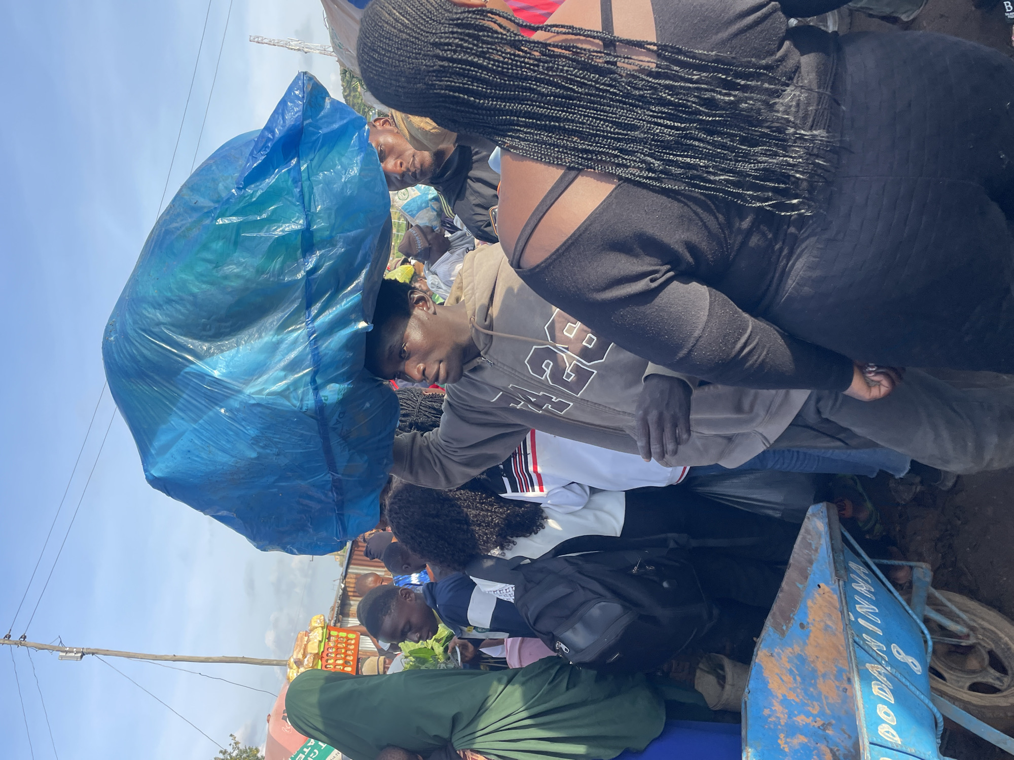 In Gura Top, Nigeria, a group of people are gathered outdoors. In the foreground, a man is carrying a large, bright blue plastic bag on his head. He is wearing a gray hooded sweatshirt with "LA 26" printed on it in white. To his right, a woman with long, braided black hair is wearing a black top and dark pants. Behind them, other individuals can be seen, some carrying backpacks and others wearing various clothing. A blue wheelbarrow with orange rust marks and the inscription "DO DANINNA 8" is visible on the left side of the image. The environment appears to be a bustling marketplace or street, with structures and a power pole in the background under a bright sky.