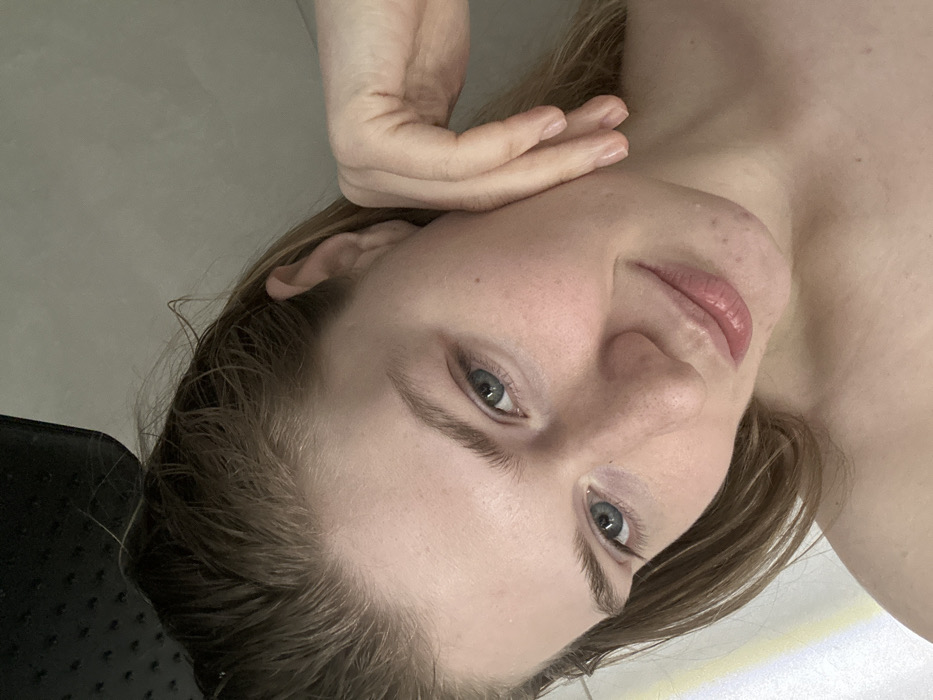 A young woman with fair skin and wet blonde hair looks directly at the camera. Her face is turned slightly to the side, and her hand rests against her cheek. She has light eyebrows and blue eyes, with a slightly upturned nose and pink lips. The background is a neutral, possibly white or grey, surface. The lighting is soft, emphasizing her features. Part of a dark, possibly textured, object is visible in the lower left corner.