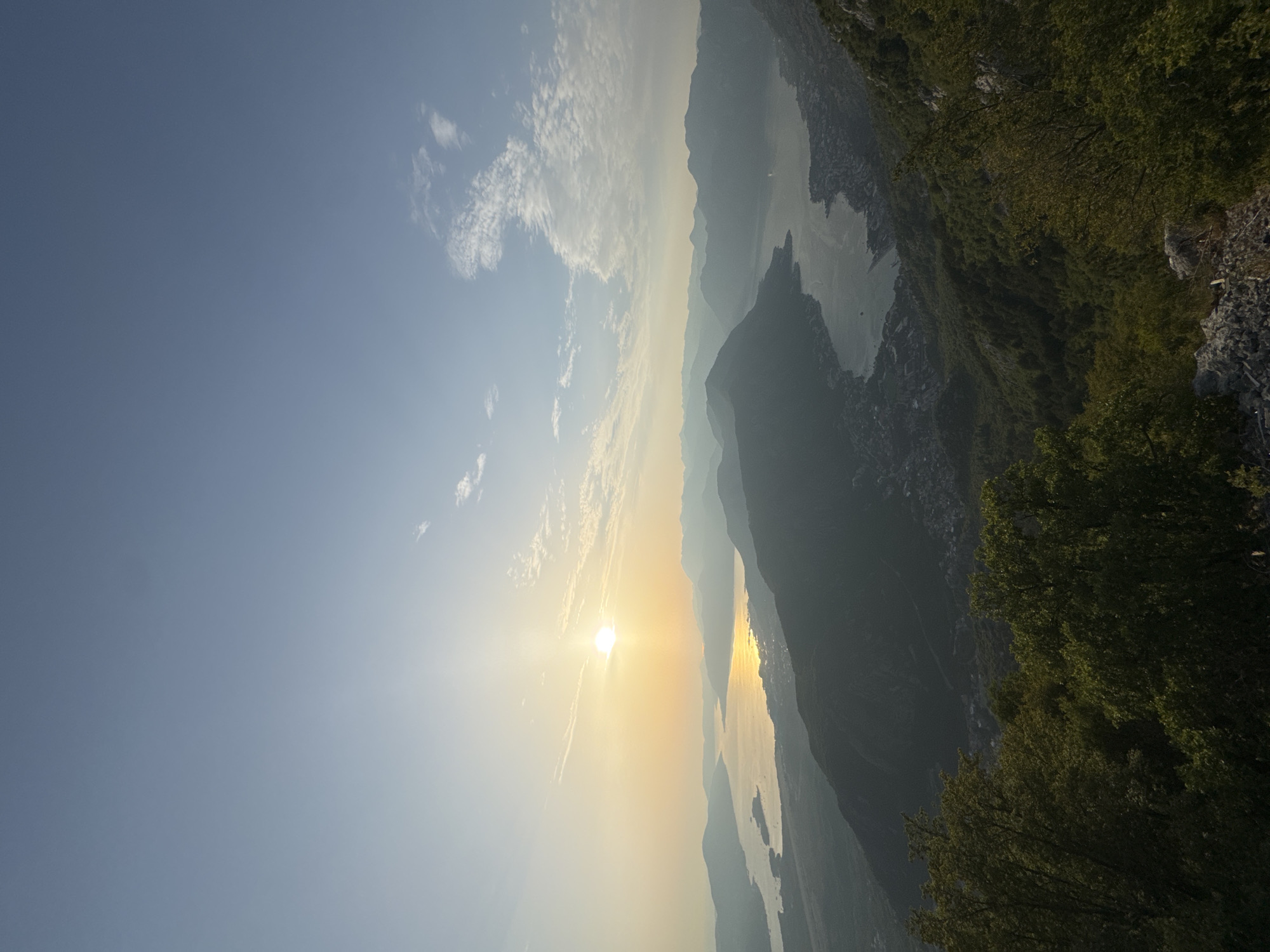 The image depicts a panoramic view of the Bay of Kotor in Špiljari, Montenegro, at sunset. The sun, a bright orb casting a warm golden glow, is positioned low in the sky towards the left of the frame, illuminating the tranquil waters of the bay. A series of layered, silhouetted mountains recede into the distance, their peaks fading into a soft haze. The bay itself is dotted with small islands and inlets, and a coastal town, with buildings clustered along the shoreline, is visible nestled at the foot of the mountains. In the foreground, lush green trees and rocky terrain provide a natural frame for the expansive vista. The sky is a gradient of pale blue, with wispy white clouds scattered across it, further enhancing the serene atmosphere of the twilight scene. There are no people or animals visible in the image.