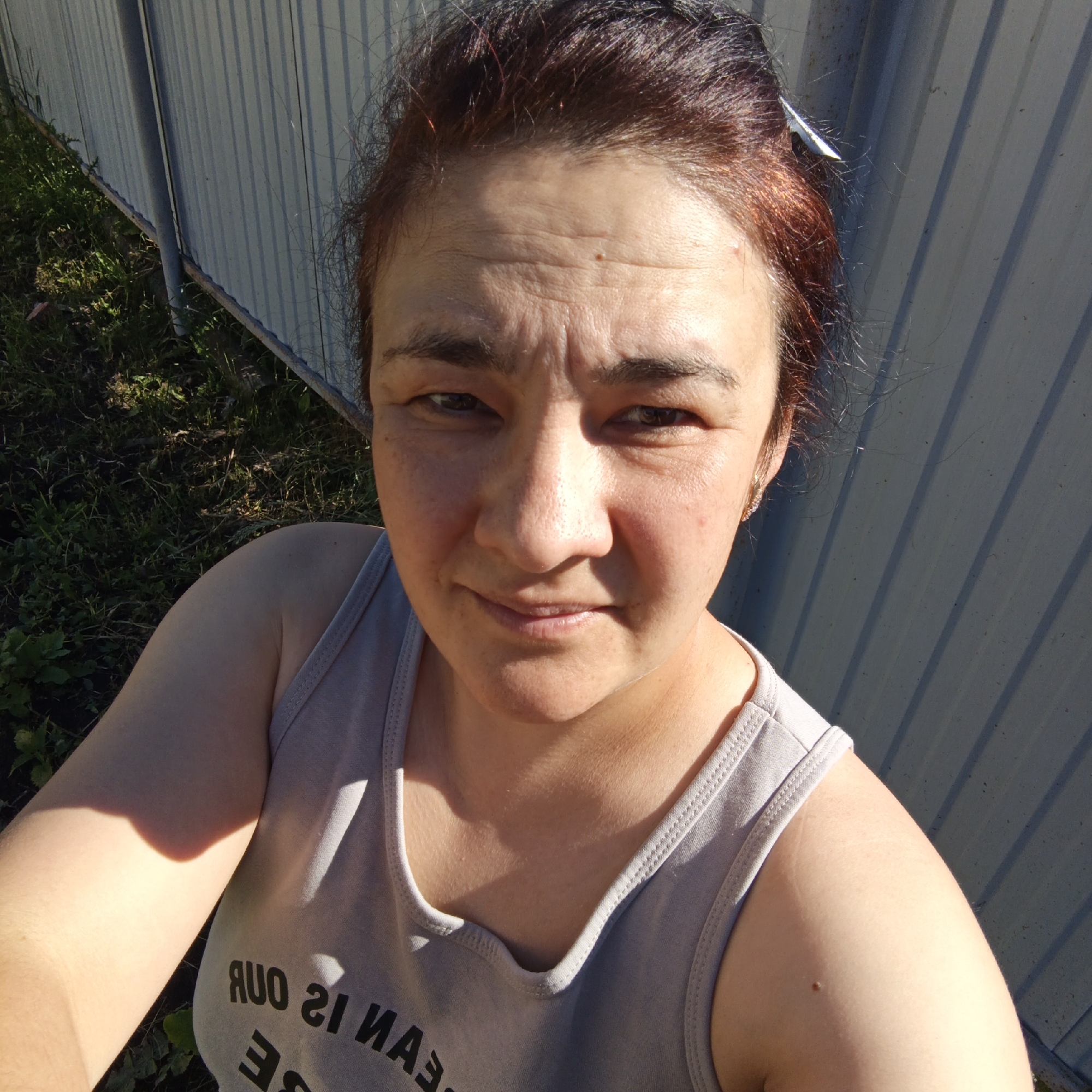 The image is a selfie of a woman in Podlubovo, Russia. She has auburn hair and is wearing a gray tank top. The background consists of a corrugated metal fence and green grass. The woman is looking directly at the camera with a neutral expression. The lighting suggests it is daytime.
