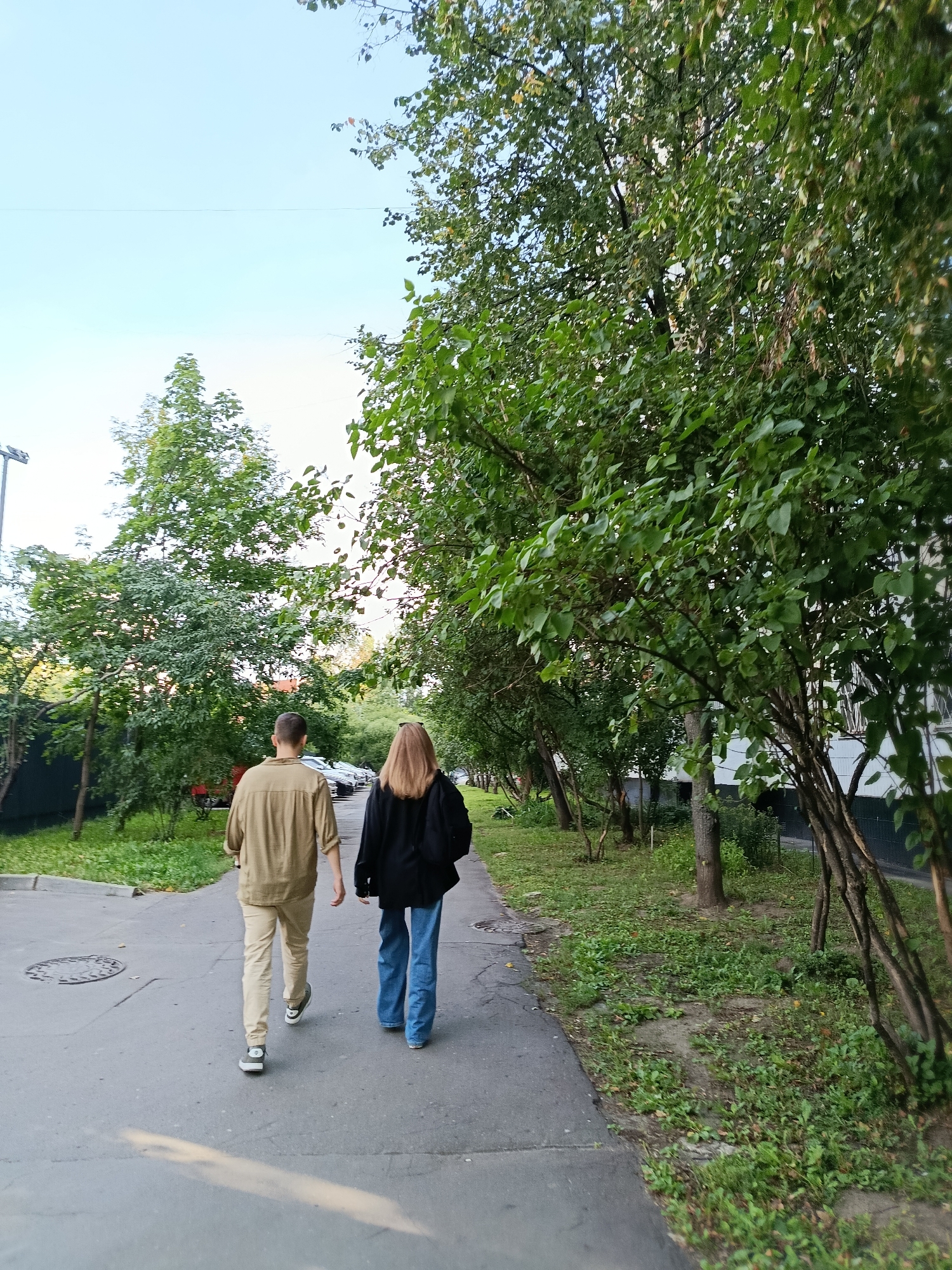 Two individuals, a male and a female, walk away from the camera on a paved pathway. The male is on the left, wearing a light brown long-sleeved shirt and light tan pants, with white and gray sneakers. The female is on the right, wearing a black hooded sweatshirt and blue jeans, with dark-colored sneakers. They are walking on a pathway lined with trees and grass on both sides. To the left of the pathway, there are parked cars and a building. The sky above is clear and blue, with some clouds. The scene appears to be in an urban residential area. Based on the architecture and vegetation, the location is likely a courtyard in a residential district in Moscow, Russia.