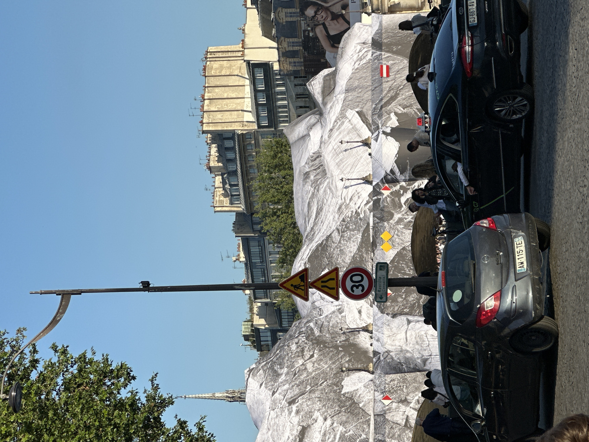 The image captures a street scene in Paris, France, showing building renovations covered in a large sheet and construction zones. There are two dark cars parked along the street. The street signs, including a speed limit of 30 kilometers per hour and directions to Bastille, confirm the location. People are visible near the construction area, suggesting pedestrian traffic. The buildings in the background are characteristic of Parisian architecture. The sky is clear blue, indicating fair weather.