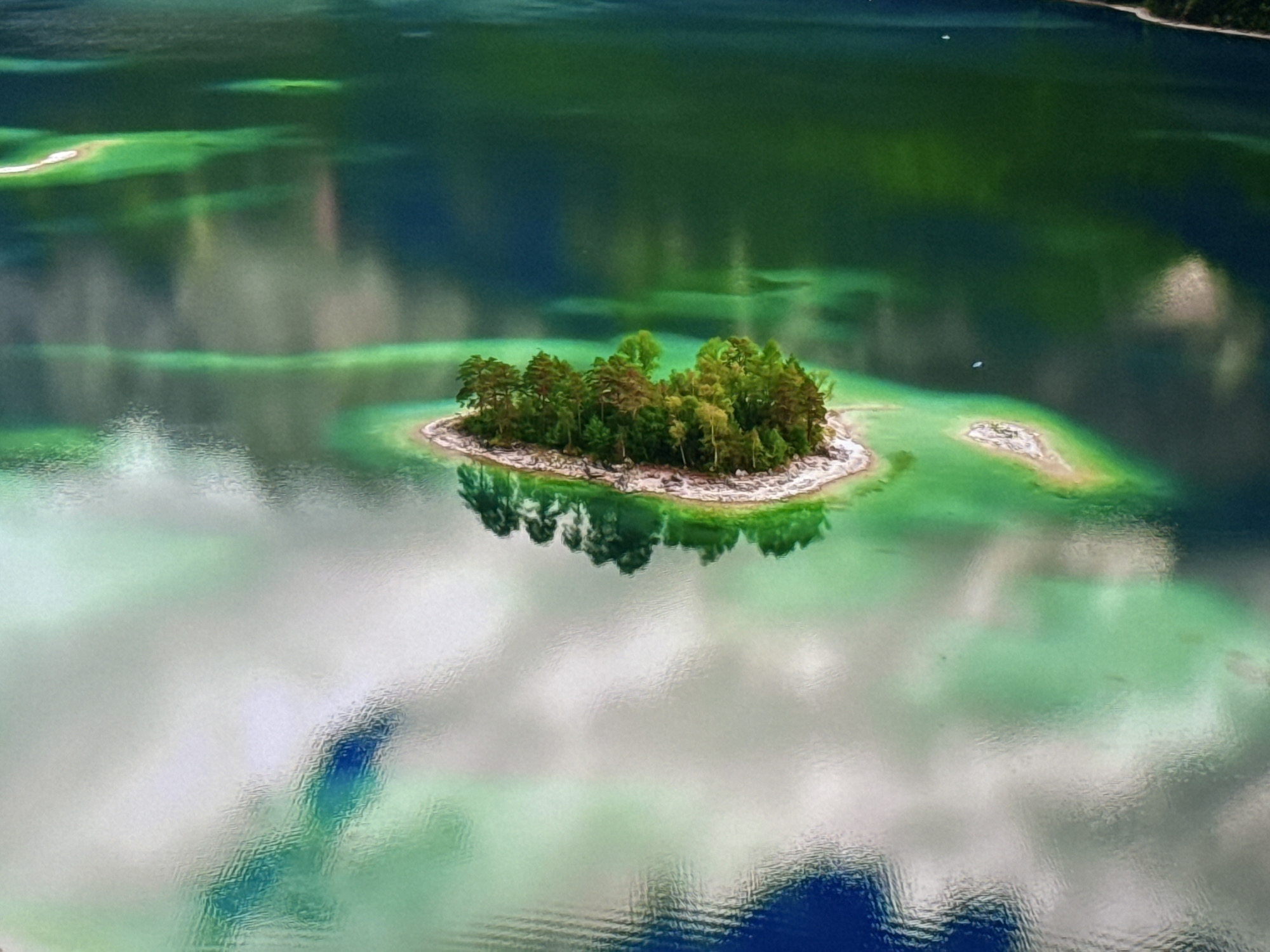 An aerial, high-angle, wide shot captures a serene, green lake with a small, tree-covered island in the center. The water exhibits a vibrant emerald hue with lighter green shallows and darker, deep blue sections. Clouds are reflected on the water's surface, creating a soft, white and gray mosaic. The island is densely populated with various trees, their reflections shimmering on the water. In the distance, other small landmasses are visible, and faint reflections suggest the presence of boats on the water. The overall scene is one of natural beauty and tranquility. This is a view of the Atanasovsko Lake in Burgas, Bulgaria.