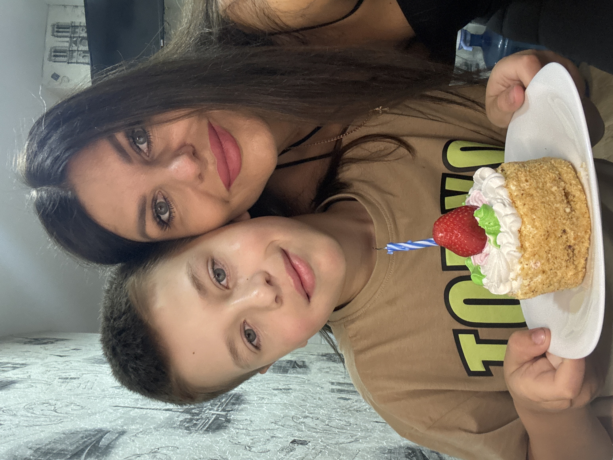 A medium shot captures a young boy and a woman posing for a photograph with a small cake in the foreground. The boy, positioned on the left side of the frame, is wearing a tan t-shirt with a lime green graphic. He holds a white plate with a single-tiered cake topped with white frosting, green and pink decorations, a strawberry, and a blue and white candle. The woman, on the right, has long dark hair and is wearing a black top. They are both looking at the camera, and the woman has her hand resting on the boy's head. The background consists of a textured wallpaper with a black and white pattern, possibly depicting buildings. The overall scene appears to be indoors, suggesting a personal celebration. The image does not provide any specific clues about the exact location within the city of Chaikovsky, Russia.