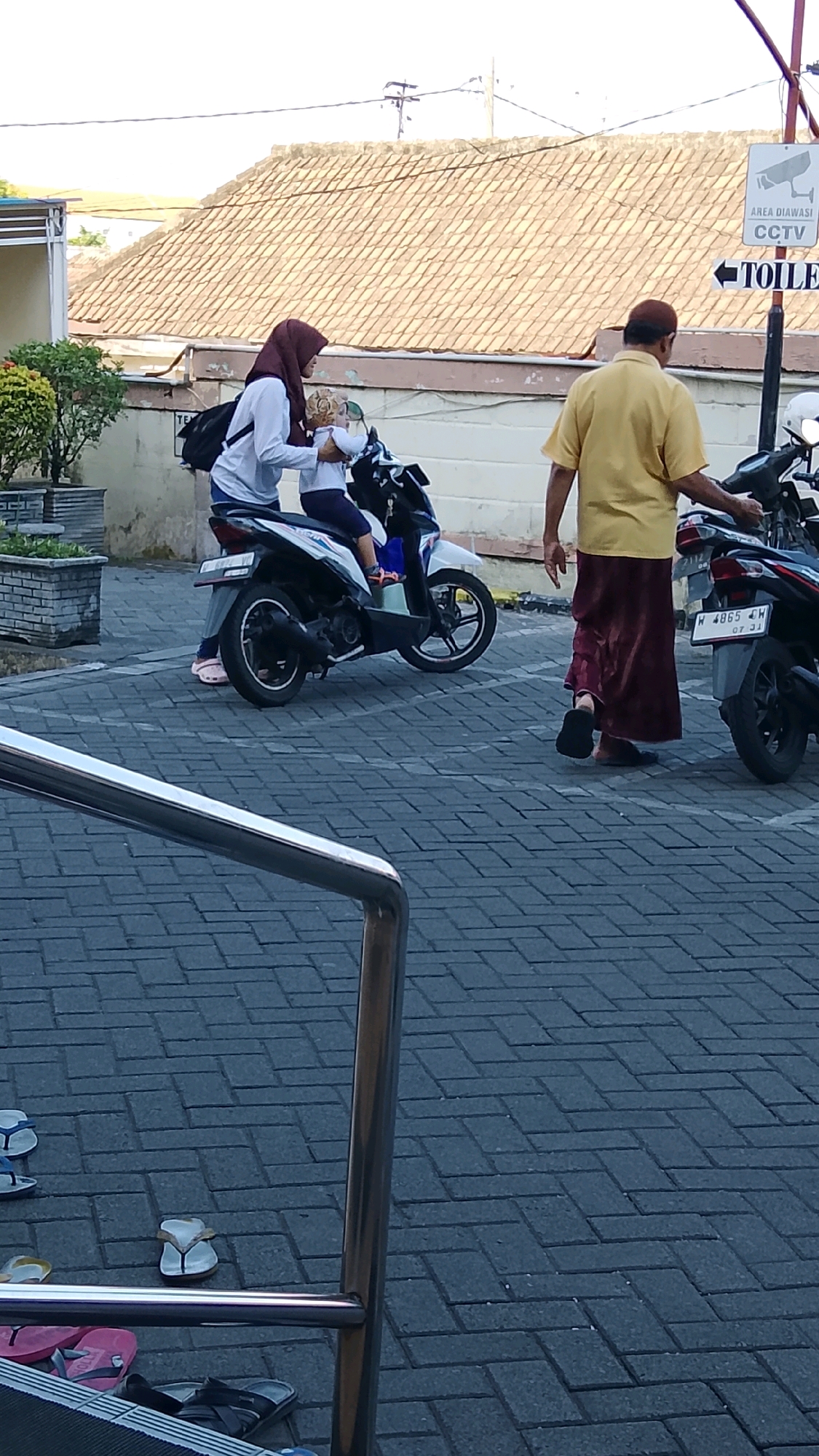 The image depicts a street scene in Ngipik, Indonesia. In the foreground, a chrome railing suggests a pathway or stairs. The ground is paved with dark grey interlocking bricks. Further back, a white and blue motorbike is parked, with a woman in a maroon hijab and white shirt sitting behind it. She is holding a child dressed in blue. To the right, a man in a yellow shirt and maroon skirt walks away from the motorbike, heading towards another parked motorbike. A sign is visible on a pole behind the man, indicating "AREA DIAWASI CCTV" (Area monitored by CCTV) and an arrow pointing to "TOILE". The background shows a building with a tiled roof. There are several pairs of flip-flops scattered on the ground in the lower left and right portions of the image.