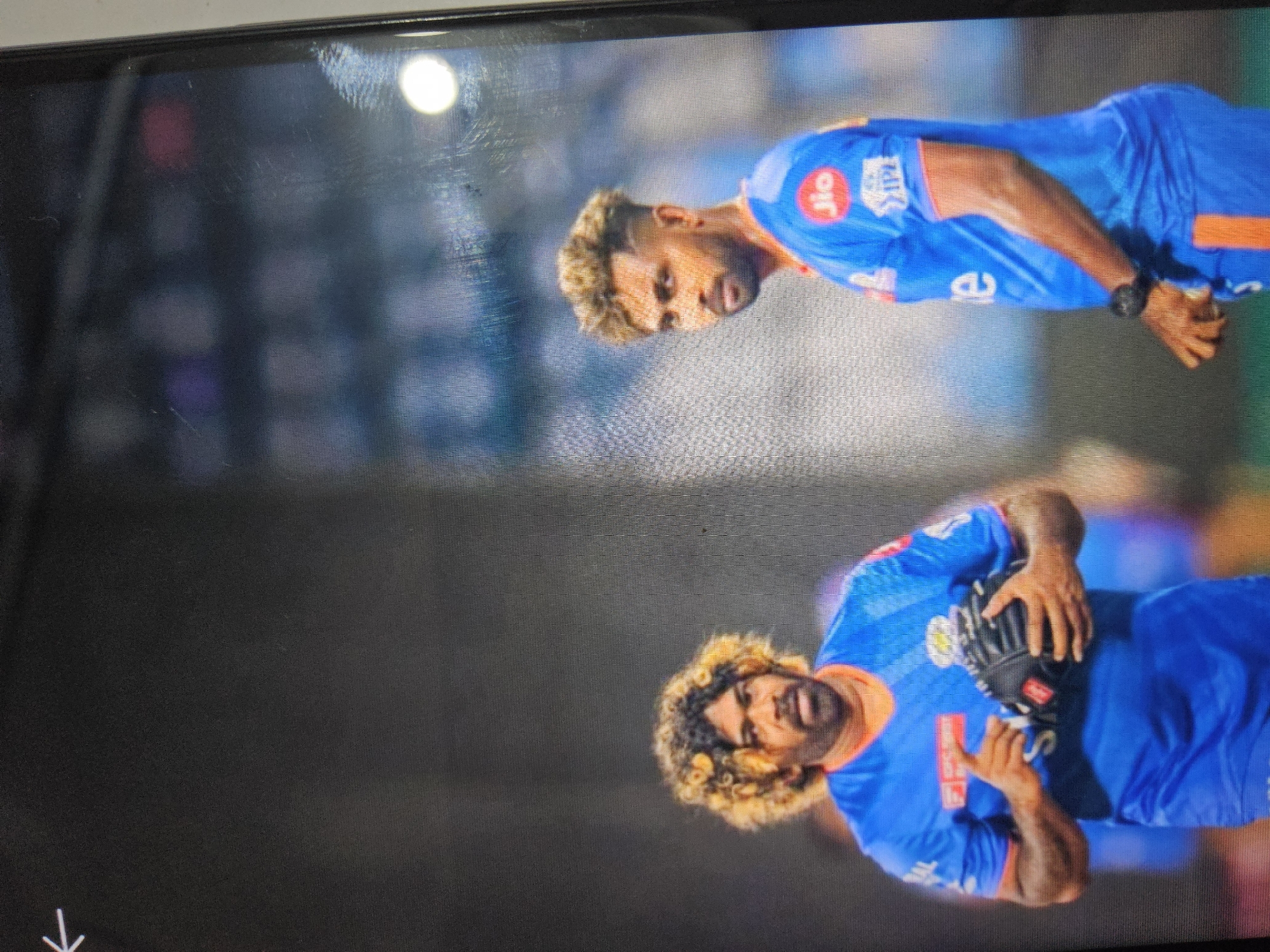 The image shows two male cricketers wearing blue jerseys with orange trim. The man on the left has a wild, curly, blonde-streaked hairstyle and is holding a black baseball glove. The man on the right has short, spiked hair and is looking towards the left. Both men appear to be on a sports field, likely during a match, with a blurred background of a stadium or crowd. The location in the city of Payagaboh, Indonesia, is not discernable from the image.