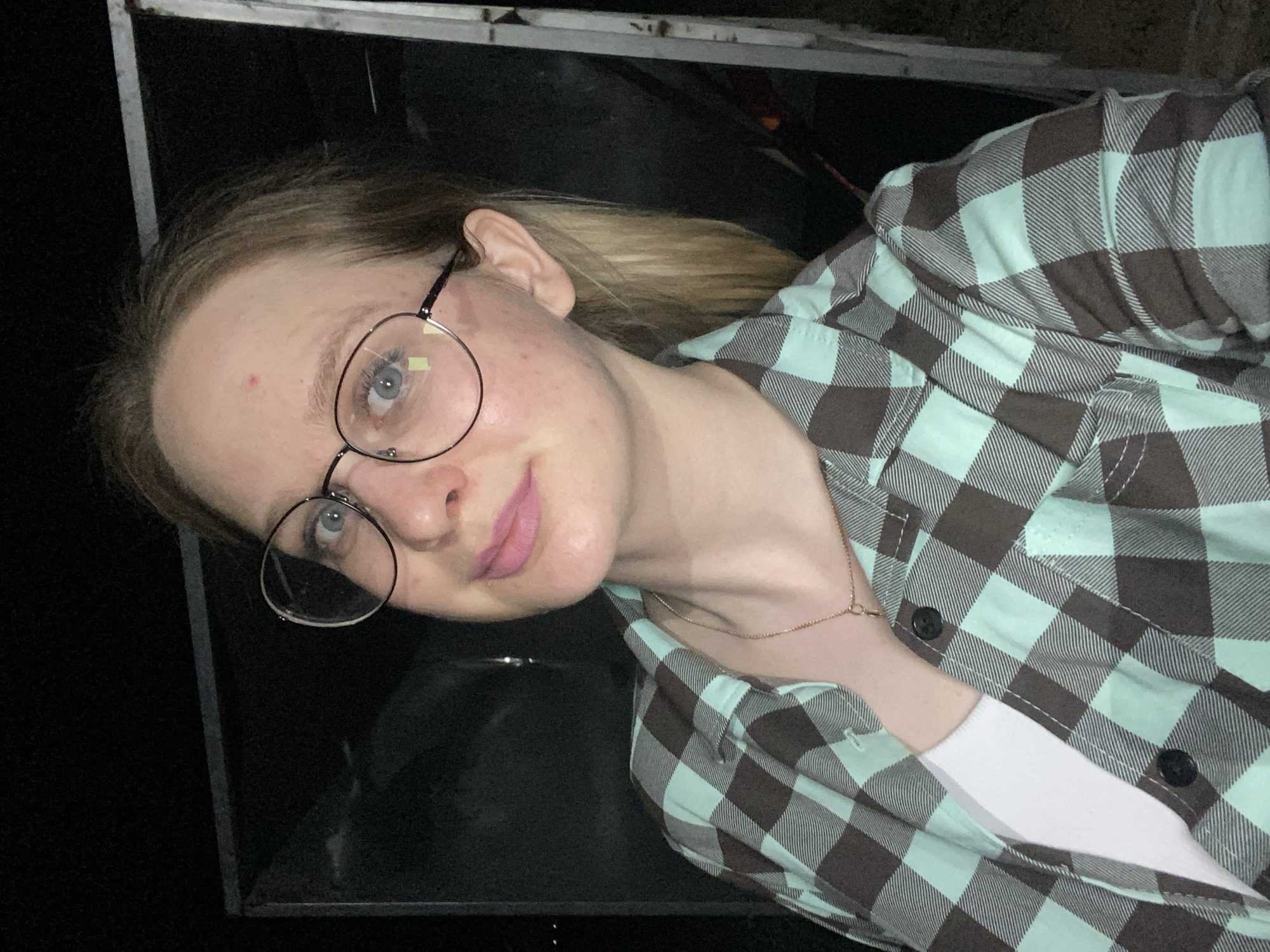 A young woman with blonde hair, fair skin, and wearing round glasses is seen indoors. She is wearing a light green and dark brown checkered flannel shirt over a white top and a delicate gold necklace. The background is dark, with a black object taking up a large portion of the frame behind her. The location is unidentified.