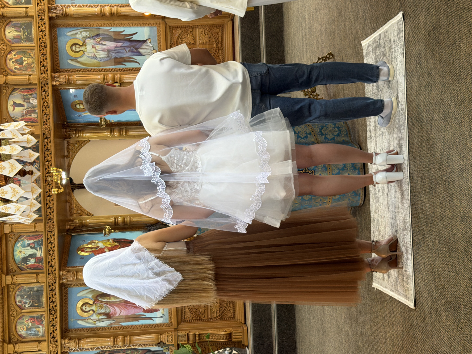 In an Orthodox church in Strășeni, Moldova, three people stand facing an iconostasis, a wall of religious icons. A woman on the left, with her back to the camera, wears a brown tulle skirt and a white headscarf. To her right, a bride in a white veil and short wedding dress stands. On the right, a man in a white t-shirt and blue jeans faces the iconostasis. The scene is illuminated by ambient light, highlighting the ornate details of the church's interior, including icons and carved wood.