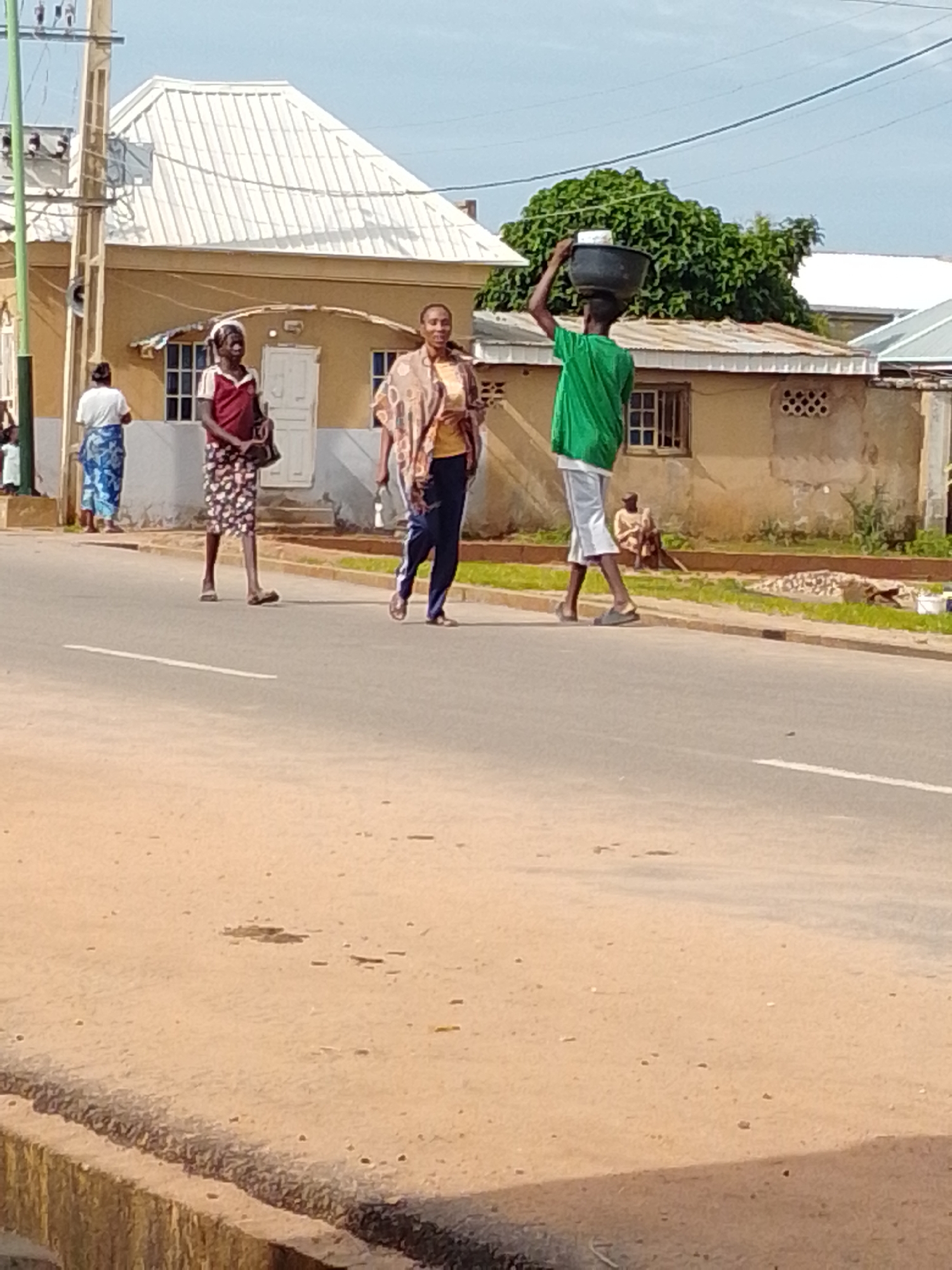 In Gombe, Nigeria, a man in a green t-shirt carries a dark basin on his head while walking on a paved road. To his left, a woman in a patterned dress and a headscarf walks away from the camera, holding a bag. Another woman in a white shirt and a blue and white patterned wrap walks towards the camera. Behind them, a building with a white roof and yellow walls is visible, with a large green tree to the right. The scene appears to be a street in a residential area.