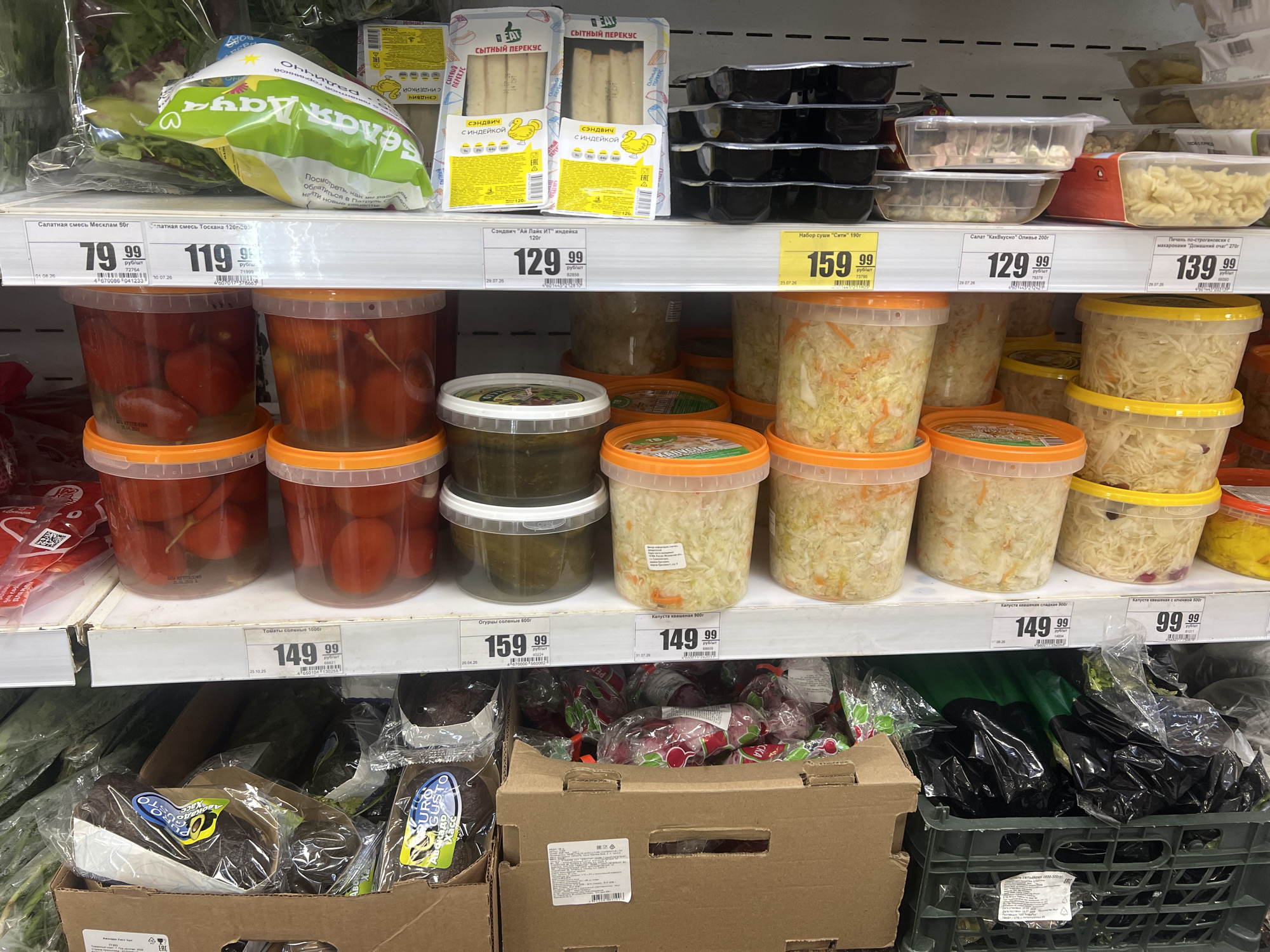 The image shows a retail store shelf displaying various food items. In the foreground, on a lower shelf, are several large plastic containers filled with what appears to be pickled tomatoes and sauerkraut. Above these, on a higher shelf, are more containers of sauerkraut and other pickled vegetables. On the shelf above that, there are packaged sandwiches and black plastic containers. Further up, there is a bag of salad greens. The shelves are made of white material, and yellow price tags are visible throughout the display. The lighting suggests an indoor retail environment. The products are predominantly packaged in plastic containers and bags. There are no people visible in the image.