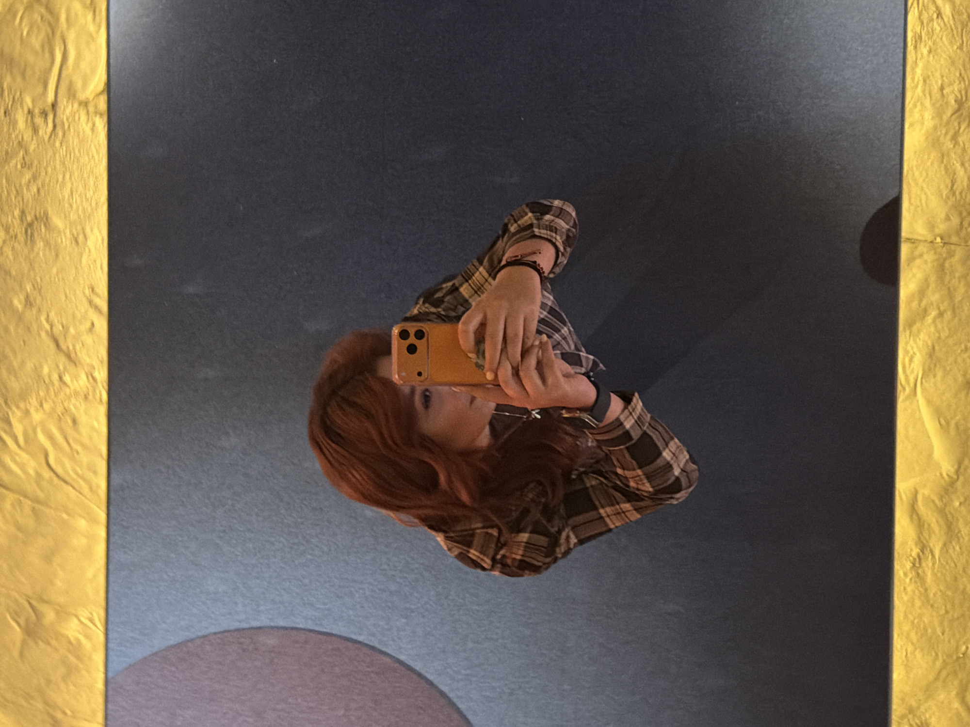 A selfie of a person is taken in a room with an abstract painted floor and textured gold-painted walls. The floor consists of a blue-grey surface with a partial pink circle. The person, with long red hair, is wearing a plaid shirt and holding a yellow phone. The location is Yerevan, Armenia.
