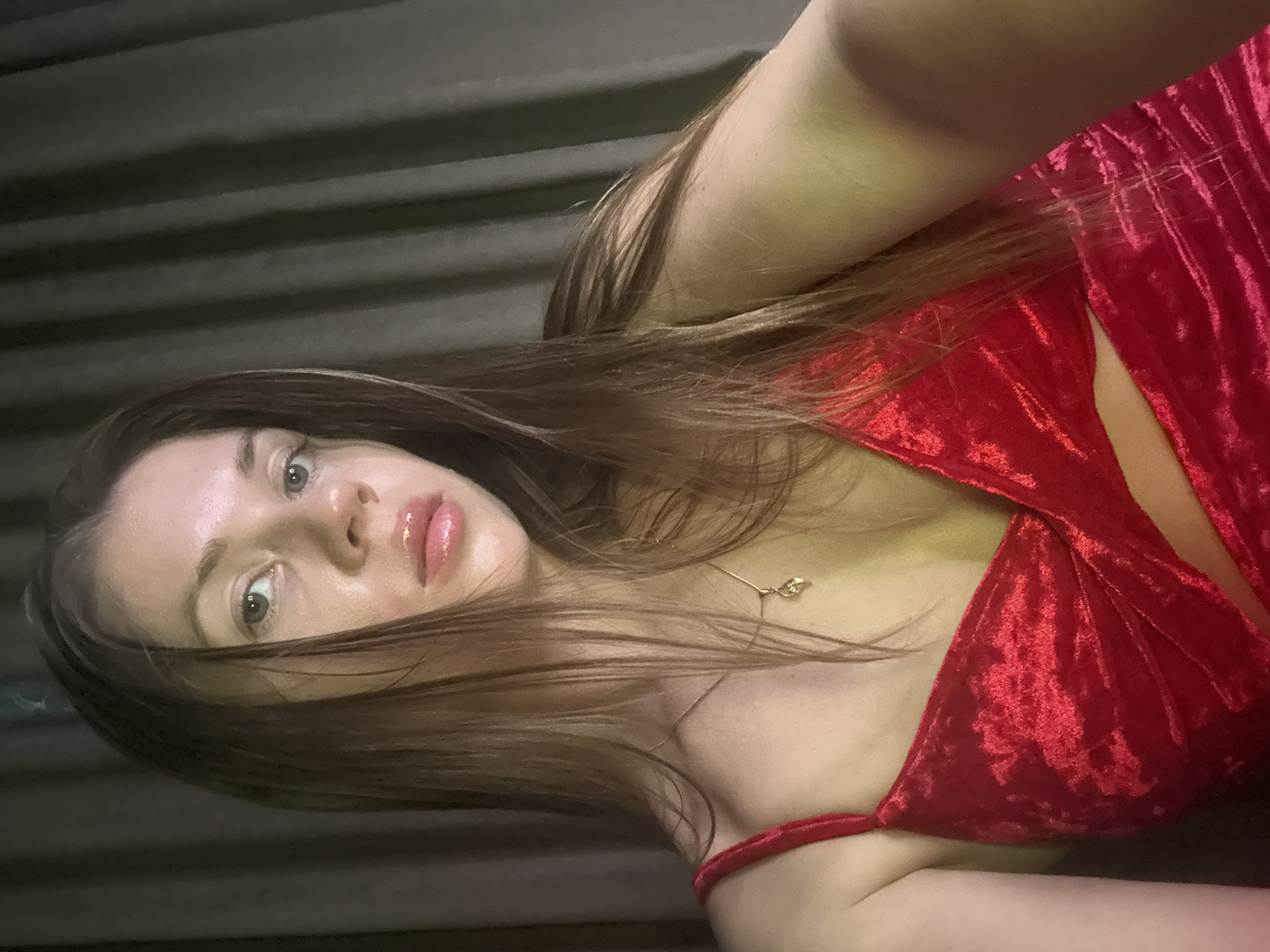 A woman with long brown hair is wearing a red velvet dress and a delicate gold necklace. She is looking directly at the camera with a slightly tilted head. The background is a dark, corrugated metal surface. It is not possible to determine the location in Batumi, Georgia from this image.