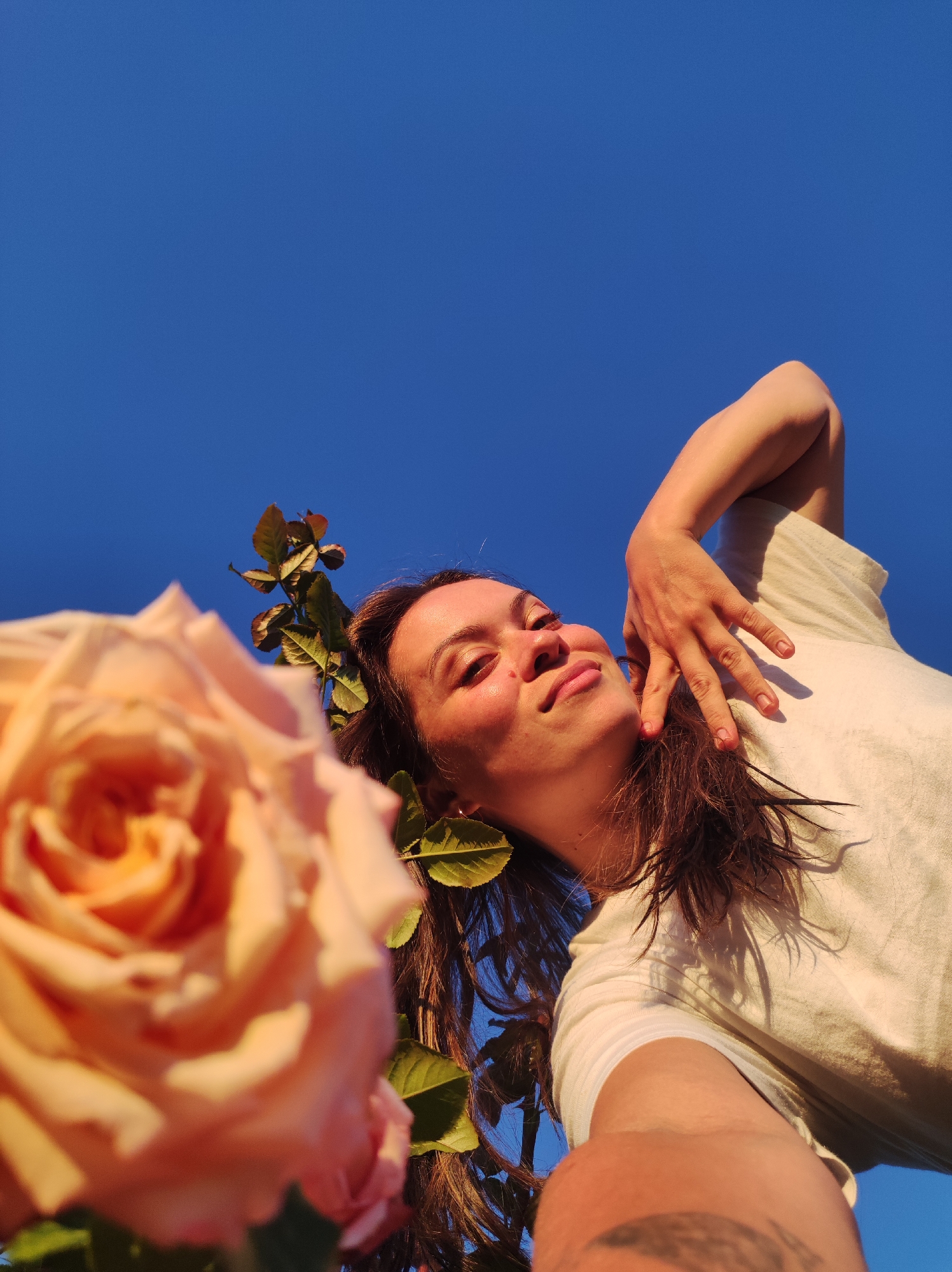 A low-angle, medium shot captures a woman with long, brown hair and fair skin, looking up with a gentle smile against a clear blue sky. She is positioned to the right of a large, out-of-focus peach-colored rose in the foreground. Her left arm is raised and bent at the elbow, with her hand resting near her face. She wears a light-colored, loose-fitting top. The scene is illuminated by warm, golden sunlight, creating soft shadows and highlighting her features. The environment appears to be outdoors, with the sky occupying the majority of the upper frame. There is no indication of the city of Фоки, Russia within the visual information provided.