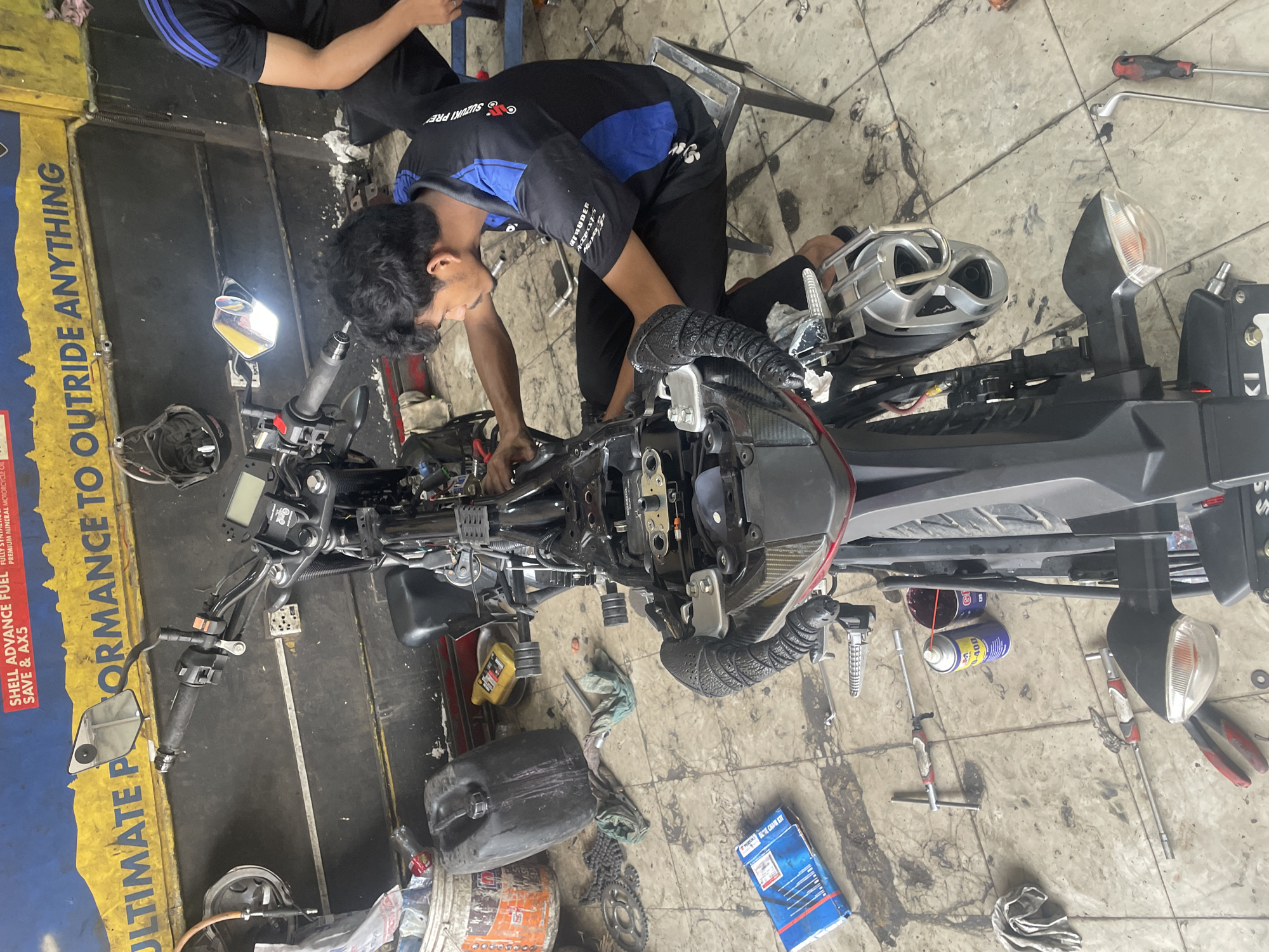 The image shows a person working on a motorcycle in a repair shop. The person is wearing a black and blue Suzuki branded shirt and black pants, leaning over the motorcycle, apparently working on the engine area. Another person is visible partially in the upper left corner. The motorcycle's body is mostly black and silver. Various tools and equipment are scattered on the floor around the motorcycle, including cans of lubricant or cleaner, wrenches, pliers, and a box labeled "Suzuki." The floor is tiled. On the left, there is a sign advertising "SHELL ADVANCE FUEL," stating "ULTIMATE PERFORMANCE TO OUTRIDE ANYTHING" and "PREMIUM MINERAL MOTORCYCLE." The overall setting suggests an automotive or motorcycle repair shop environment. The location is Dhaka, Bangladesh due to the clothing brand and Suzuki branded items.