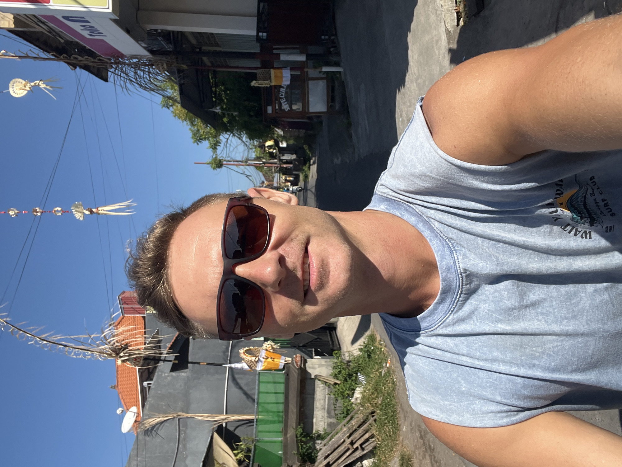 A Caucasian man wearing sunglasses and a light blue sleeveless shirt is taking a selfie outdoors. He is smiling and looking at the camera. The background shows a street in Sukawati, Indonesia, with buildings, power lines, and decorative items hanging from wires. There is a small shrine with yellow and white offerings on the left side of the image. The street appears to be paved, and there is vegetation and some wooden planks on the side of the road. The sky is clear and blue.