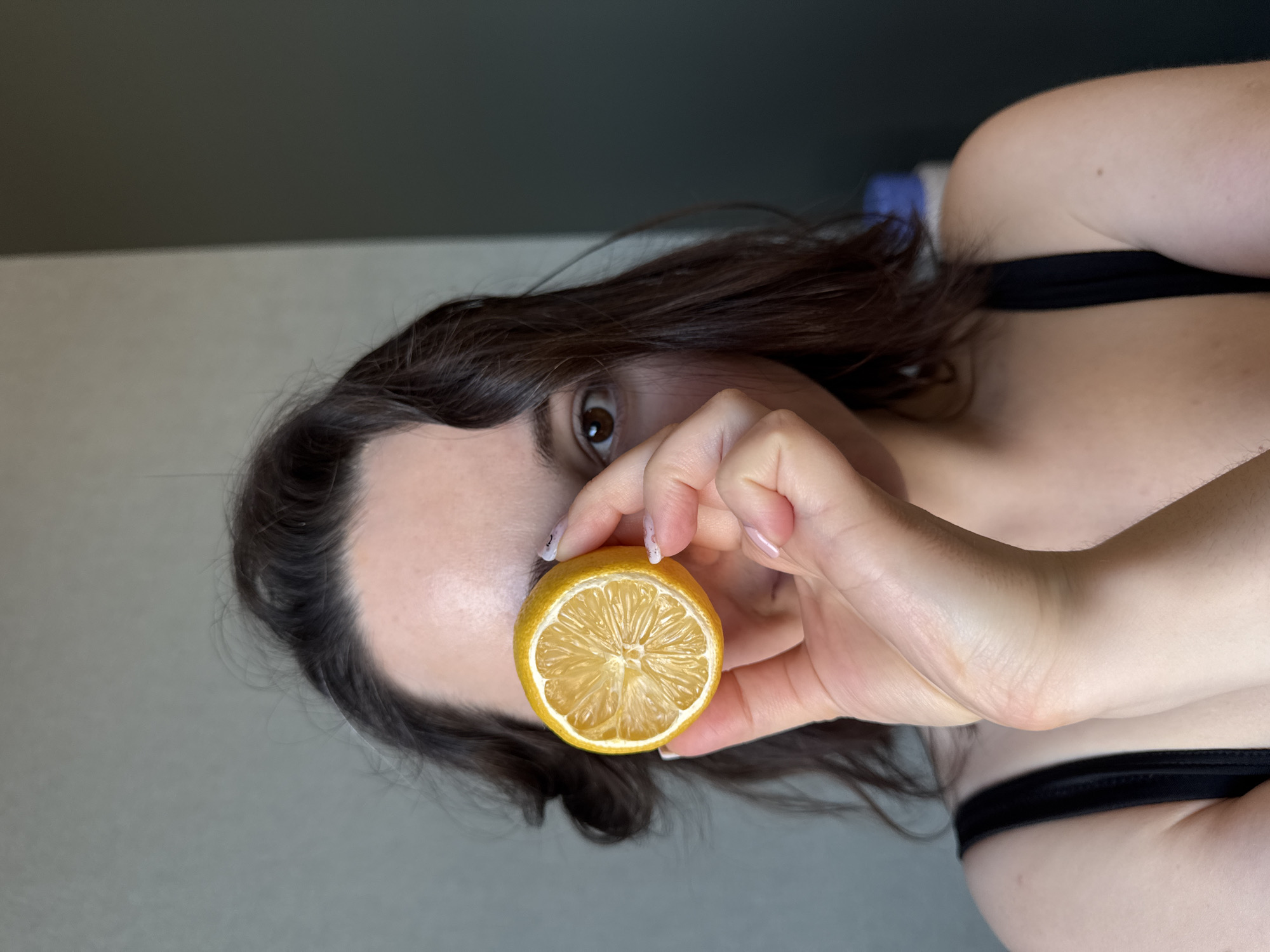 A medium close-up photograph depicts a female subject indoors. The subject, with dark brown, wavy hair, holds a dried half-lemon slice with her right hand, positioned directly over her left eye. Her right eye, brown in color, is visible and directed towards the viewer. The subject's left shoulder and part of her chest are visible, clad in a black strap garment. The background consists of a light grey wall on the left transitioning to a darker grey wall on the right, indicating an interior corner. A blue and white container is partially visible in the blurred far right background. No specific landmarks or identifying features are present to determine a precise location within Izhevsk, Russia.