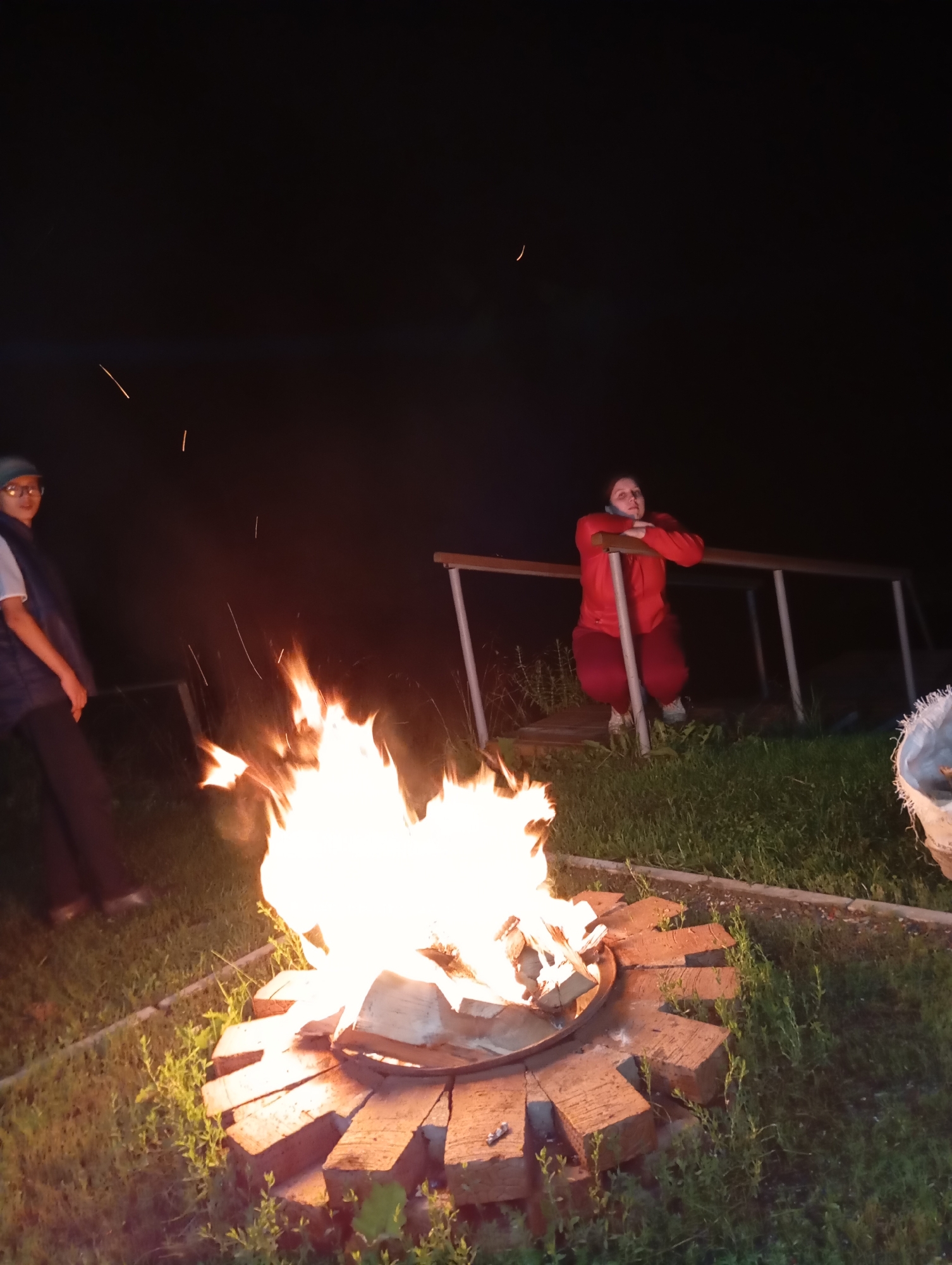 In the city of Malyi Bukor, Russia, an outdoor evening scene unfolds around a crackling bonfire. The fire, the central subject, emits bright, warm light, illuminating the surrounding grass and a circular fire pit constructed from bricks. Two individuals are present. To the left, a person wearing a dark vest over a white shirt and a green hat stands, partially obscured by the fire's glow. To the right, a person in a red hooded jacket and red pants is seated, leaning on a wooden railing. The background consists of dark, indistinct foliage and a railing. The overall atmosphere is one of a quiet, nighttime gathering.