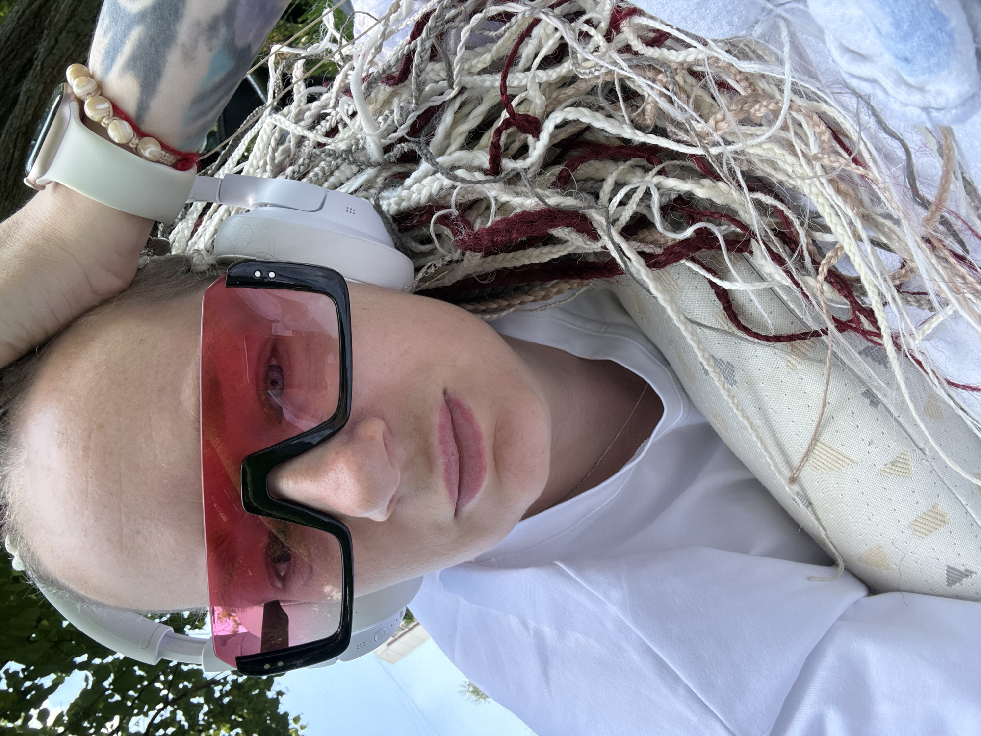 A close-up, selfie-style photograph depicts a person wearing white headphones and oversized, angular sunglasses with pink lenses. The individual has long, braided hair in various shades of blonde, grey, and burgundy. They are wearing a white t-shirt and a delicate silver necklace. A watch with a light-colored band is visible on their left wrist, along with a beaded bracelet and a red string bracelet. The background is softly blurred, showing hints of green foliage and a bright sky, suggesting an outdoor setting, potentially in Moscow, Russia. The overall impression is casual and relaxed, possibly taken on a sunny day.