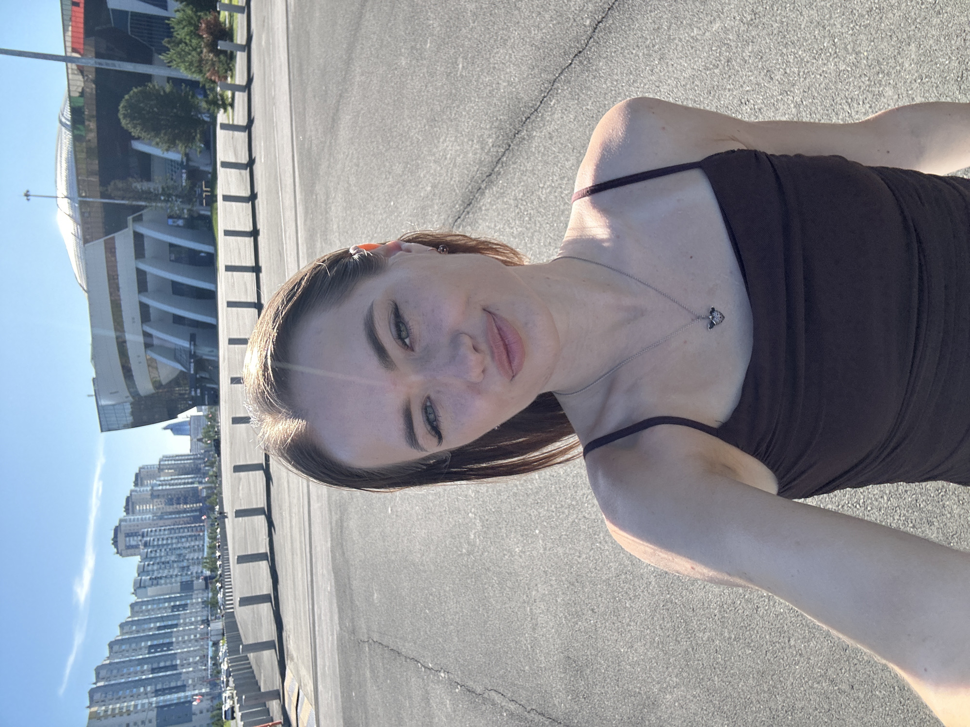A young woman with brown hair and fair skin takes a selfie outdoors. She is wearing a dark-colored tank top and a silver necklace with a small pendant. She is smiling at the camera. The background features a bright blue sky, a large modern building, and a cityscape with many tall buildings. The ground appears to be asphalt, suggesting an outdoor urban environment. The lighting suggests it is daytime, likely mid-morning or afternoon, with the sun casting shadows. The overall mood seems cheerful and relaxed. No visible text is present in the image.
