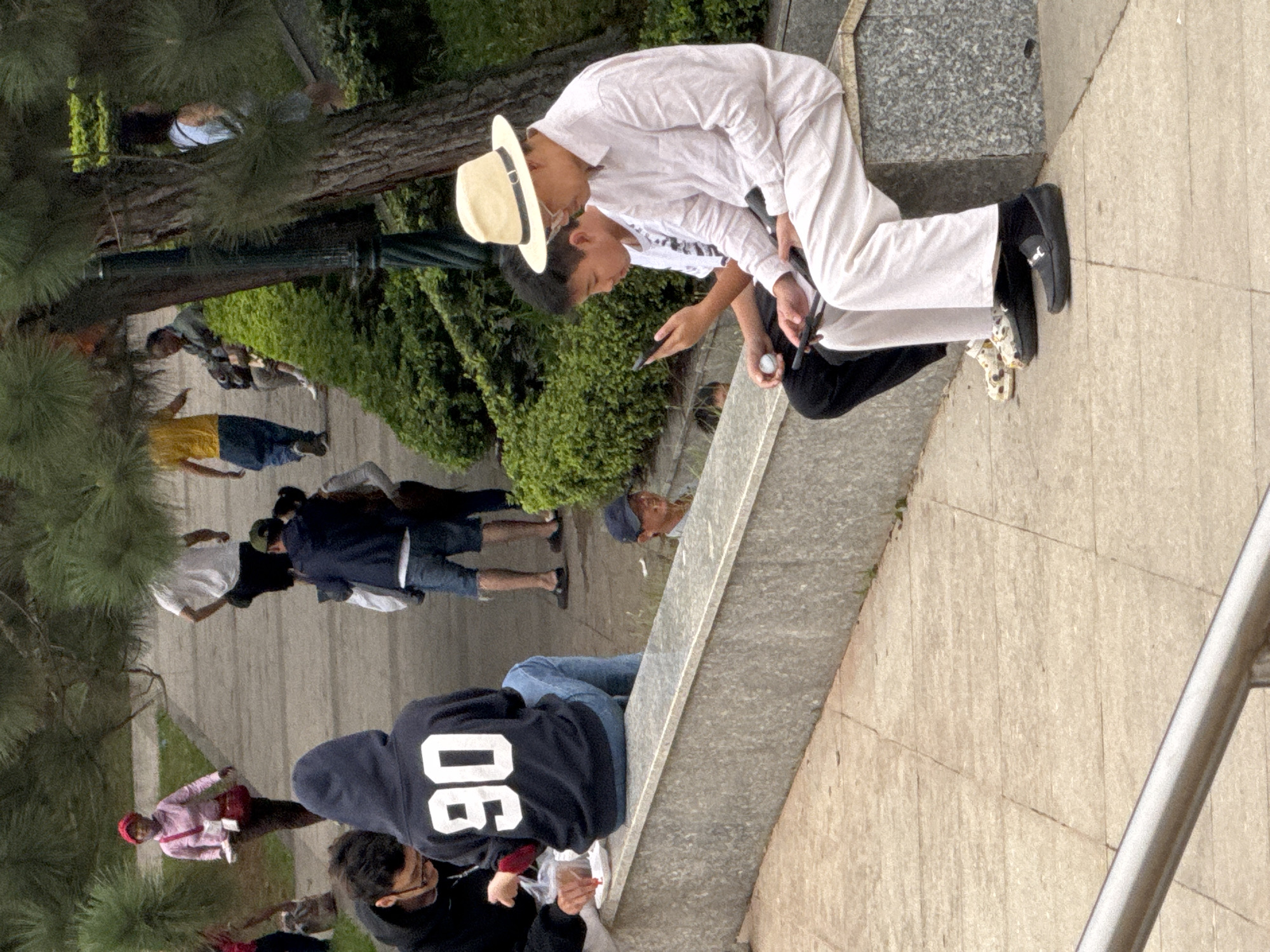 This image depicts a group of people in an outdoor setting, possibly a park or public area, given the presence of trees, a pathway, and a stone structure. The perspective is from above, looking down at an angle, and the image is rotated so that the people are oriented vertically.
The main subjects are several individuals, some of whom are standing or walking, while others are seated or interacting with each other. In the foreground, two individuals are leaning in towards each other. One is wearing a white outfit and a straw hat, and the other is wearing a white t-shirt. The person with the hat appears to be holding a phone.
Further down the pathway, other individuals are visible. One person is wearing a yellow shirt and blue shorts. Another group includes people in casual attire, including hoodies and jeans. There is a person in a pink shirt and red shorts, and another in a dark hoodie with the number "90" visible on the back.
The scene appears to be during daylight, as indicated by the shadows and the overall illumination. The weather seems pleasant, conducive to outdoor activities. The presence of greenery suggests a natural or well-maintained environment.
Notable details include the attire of the people, ranging from casual to more formal, and the various objects they are holding or interacting with, such as a phone and what looks like a golf putter. The "90" on the hoodie is a visible piece of text. The overall atmosphere seems relaxed and social, with people engaged in their own activities or interactions within the shared space. The location cues, like the greenery and the style of the pathway, are consistent with a park or garden setting.
