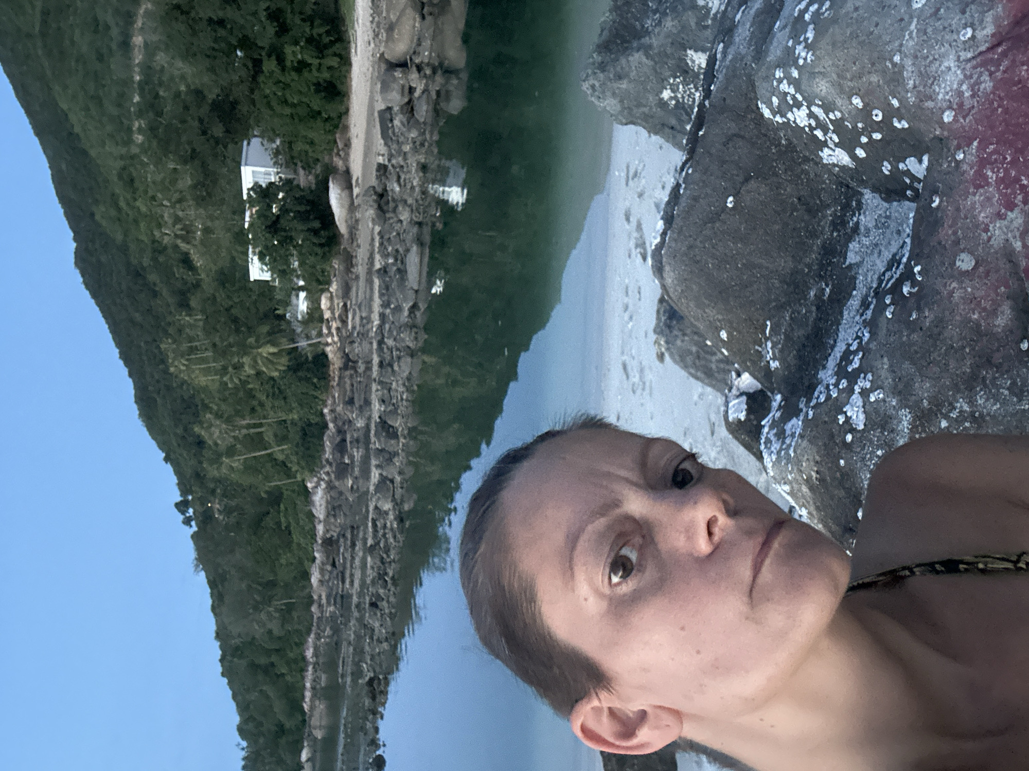 A woman is taking a selfie on a rocky beach with a calm body of water and a lush, tree-covered hill in the background. A white building with large windows is visible nestled into the hillside. The sky is a clear, pale blue, suggesting it is either early morning or late evening. The woman's face is close to the camera, and she appears to be looking directly at it. The water is still and reflects the greenery of the hill and the sky. Rocks covered in white barnacles are in the foreground. The scene is located in ตำบลเกาะพะงัน, Thailand.